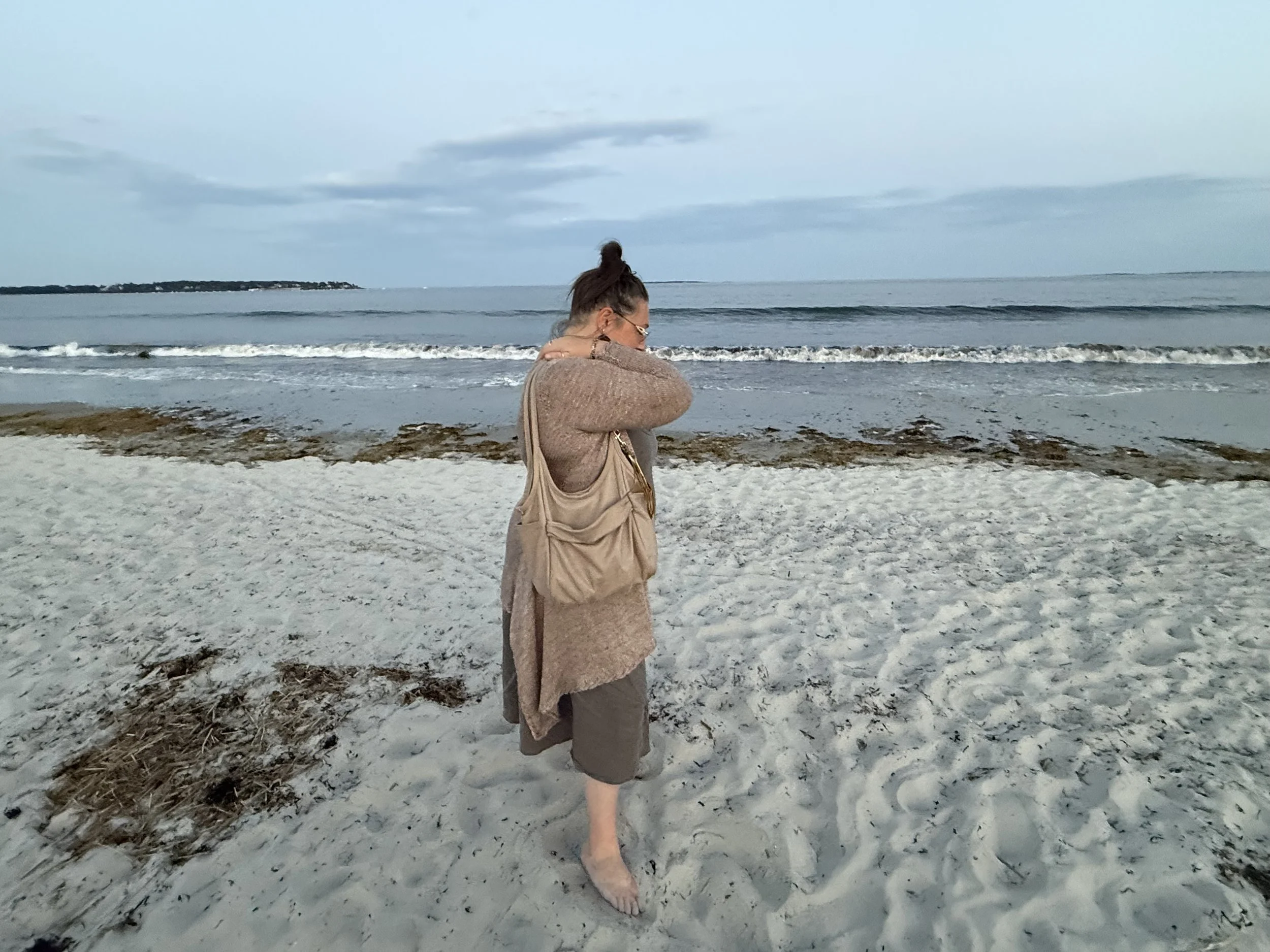 Jennifer in OOB wearing everything ATCo.- the dress, the sweater, and the gorgeous Carmel handbag standing in front of the ocean.