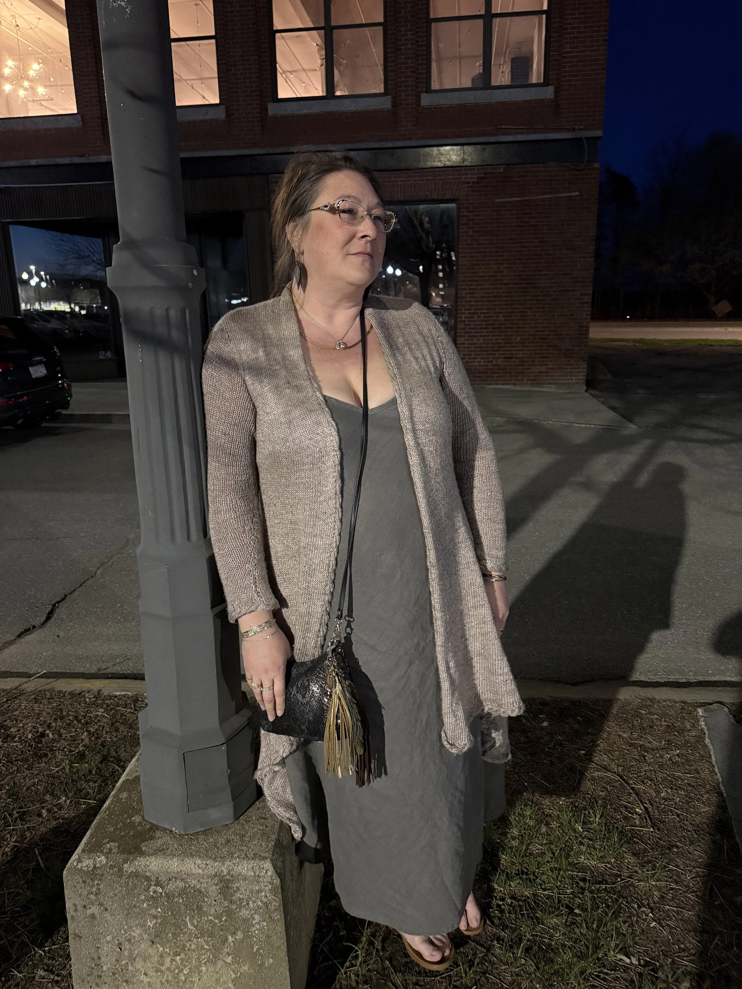 Jennifer standing outside next to a lamp post at night, wearing all ATCo., holding an Anson purse created in metallic snake, in front of a brick building with illuminated windows.
