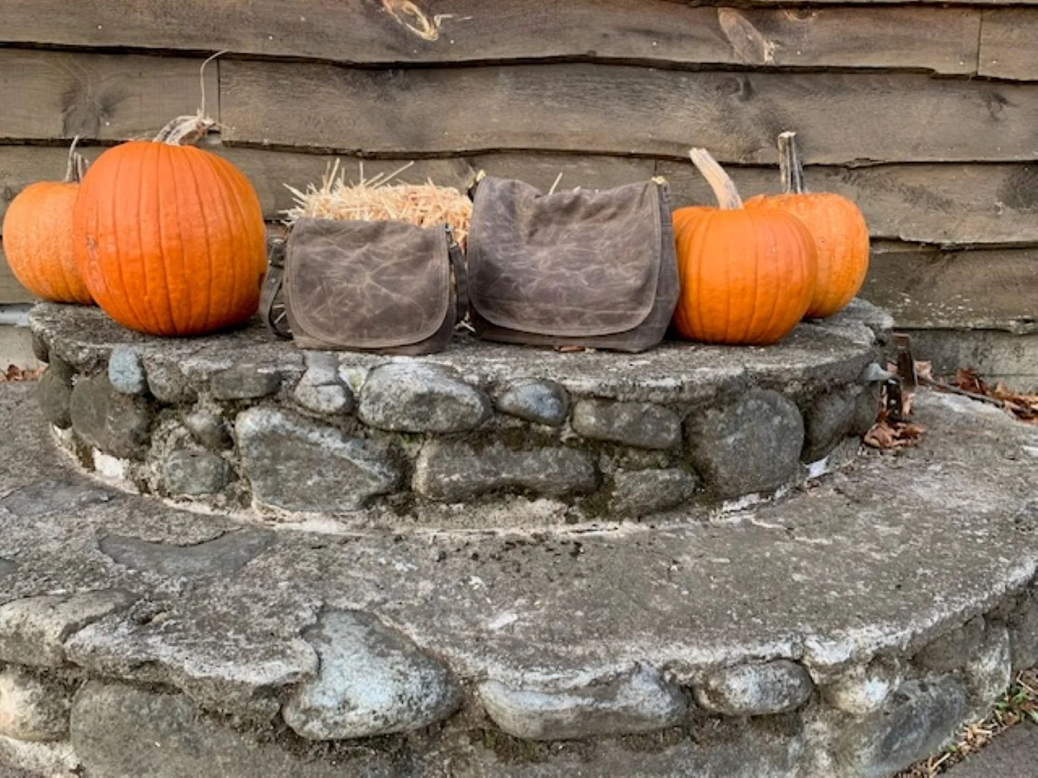 Three orange pumpkins and two canvas Eustis bags on a stone circular fire pit in front of a wooden fence.
