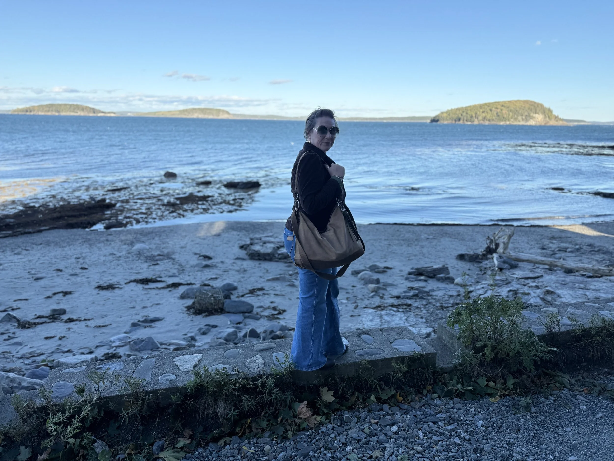 Jennifer standing on a rocky beach near the water, with islands visible in the background under a blue sky- ATCo. makes heirloom-quality leather handbags with a focus on intentional relationships with customers.
