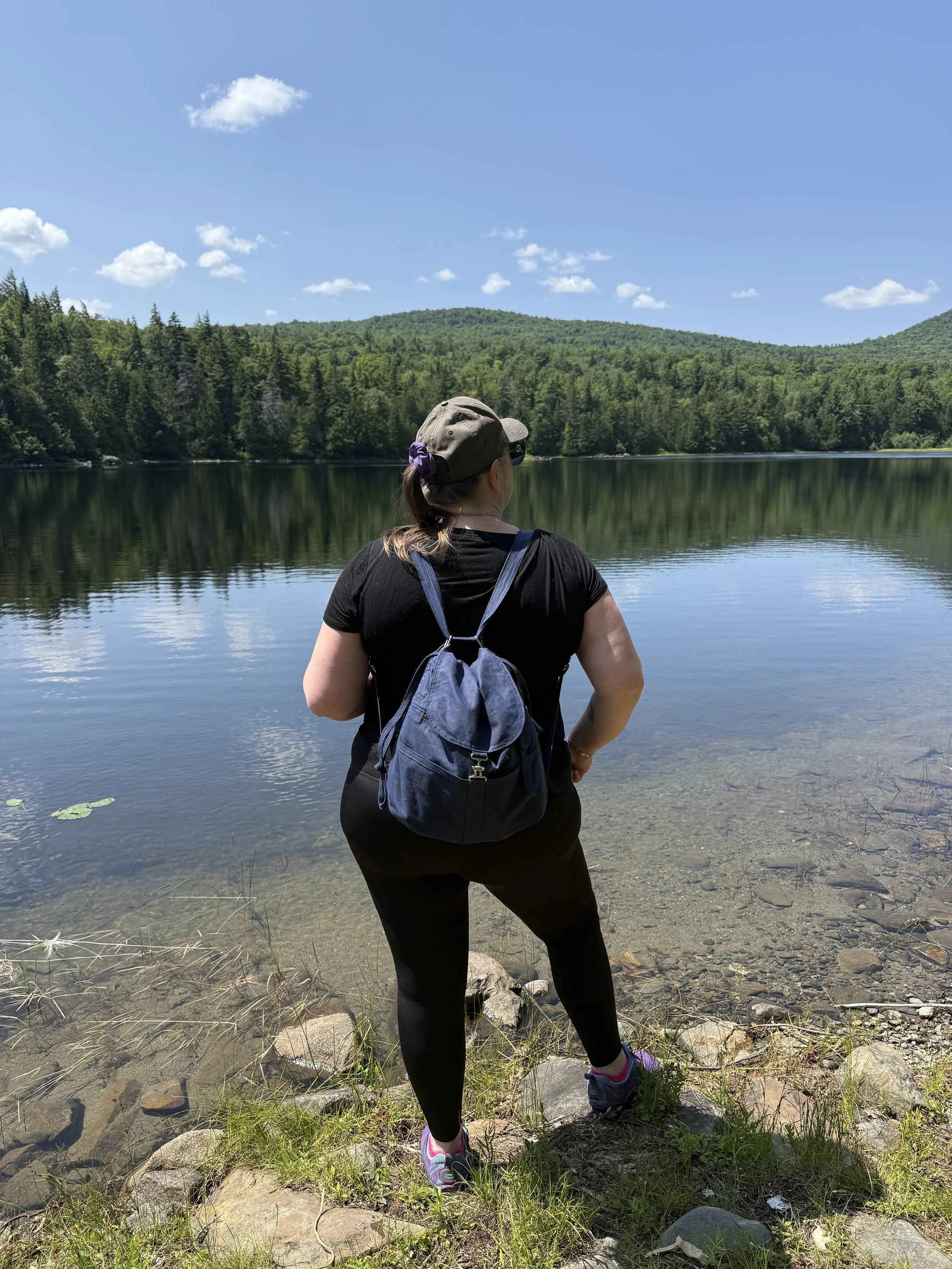 Jennifer standing on the bank of a lake, facing away, with the Appalachian Mountains and a blue sky with clouds in the background.