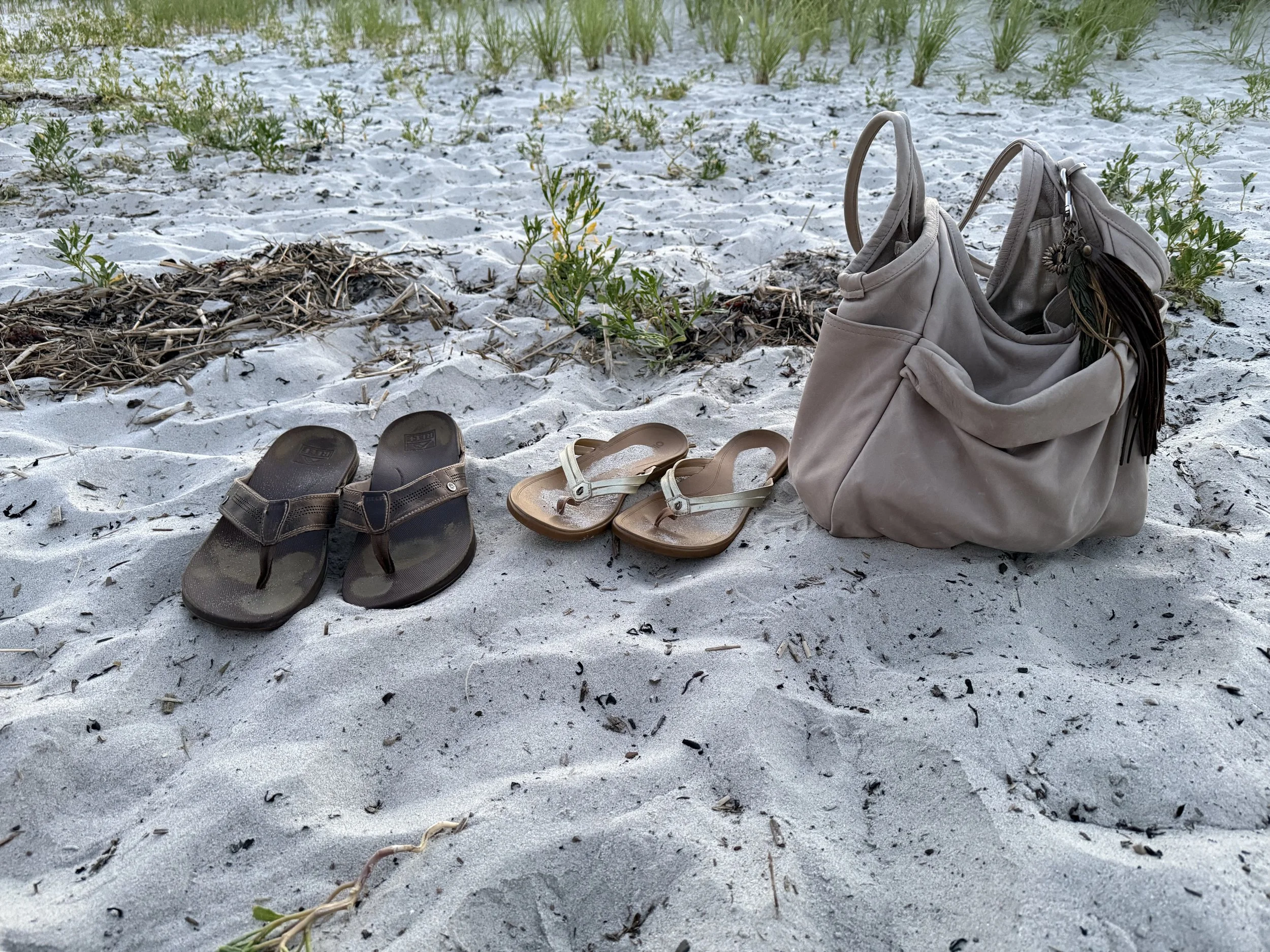 A sandy beach with some green plants emerging from the sand. There are two pairs of sandals, one black and one tan, and the Carmel handbag with bag bling hanging from it.