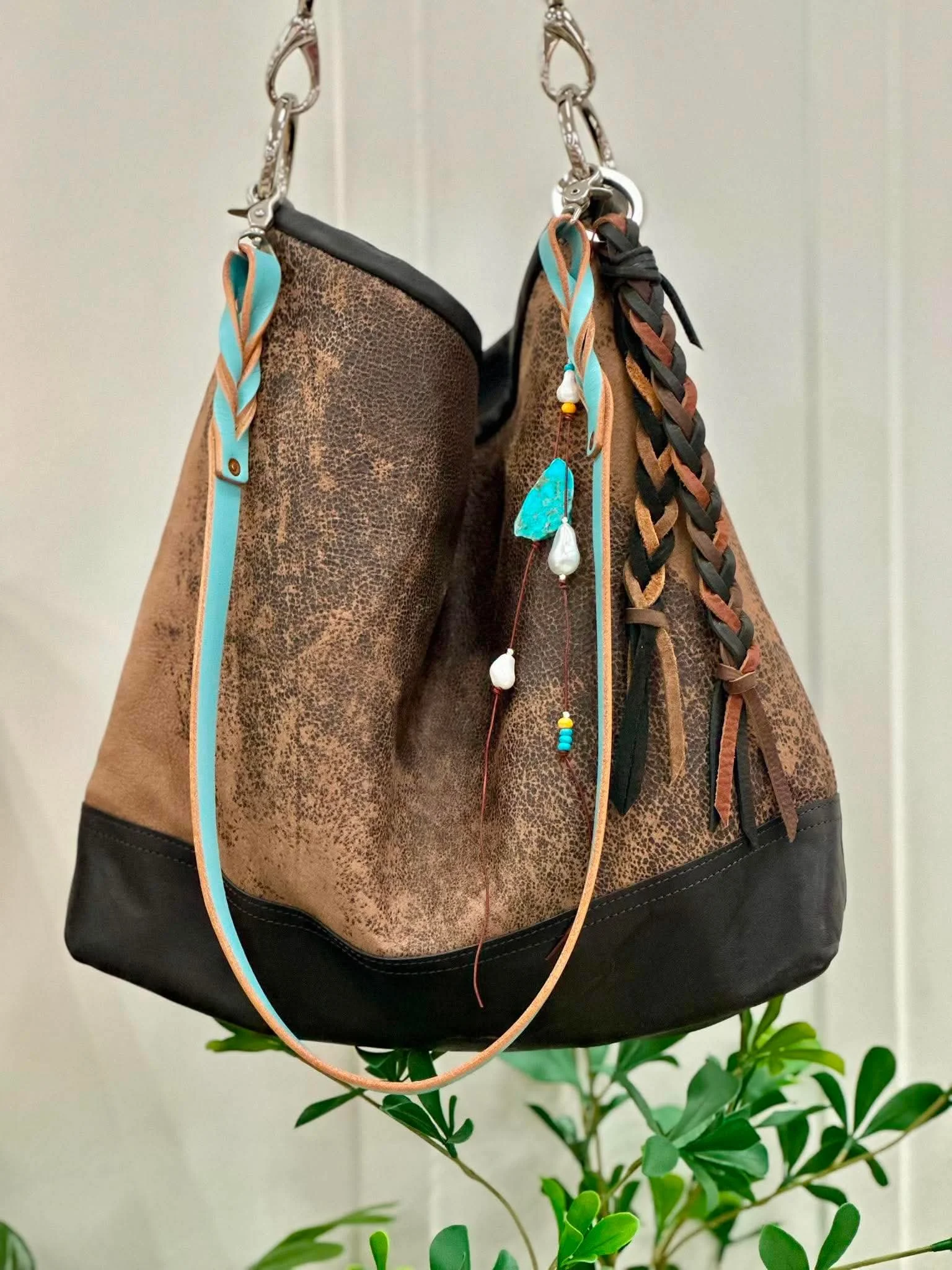 A bespoke, custom Bar Harbor hanging on a hook, decorated with small colorful beads and a turquoise ribbon. The bag is hanging in front of a potted plant with green leaves, against a light-colored wall.