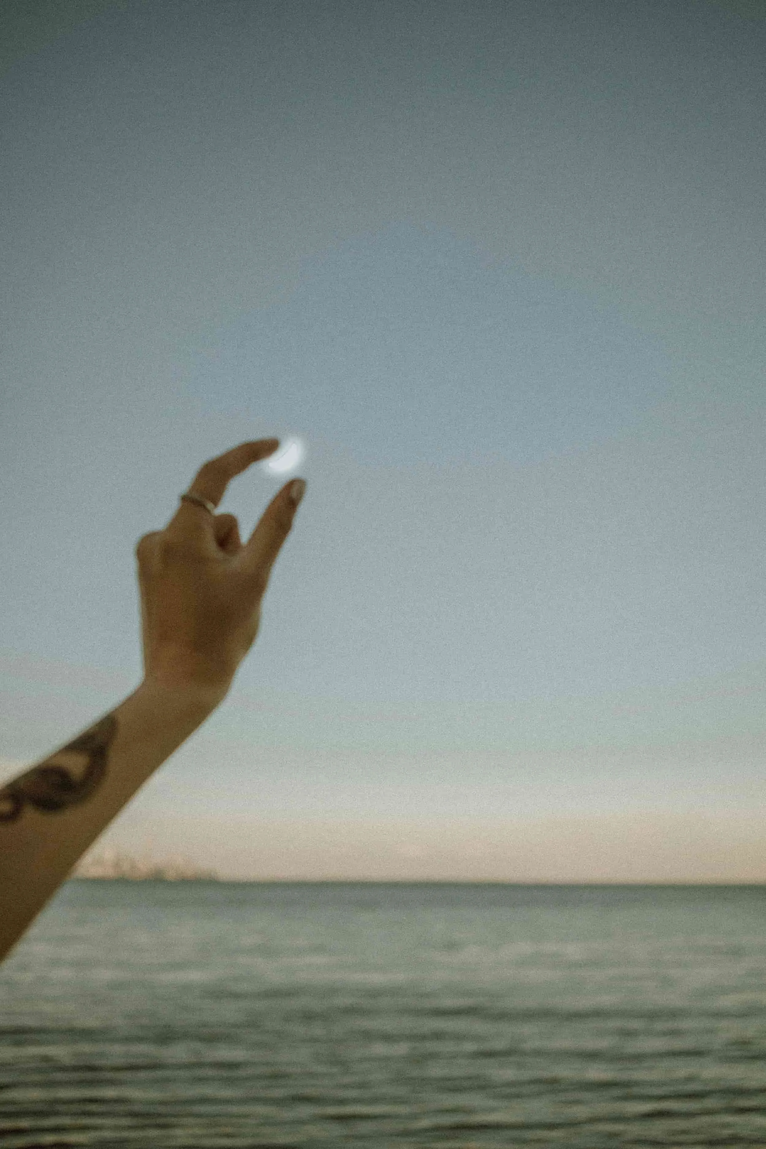 A person's hand with tattoos and rings, holding a small crescent moon-shaped object against a sky and body of water at sunset or dusk.