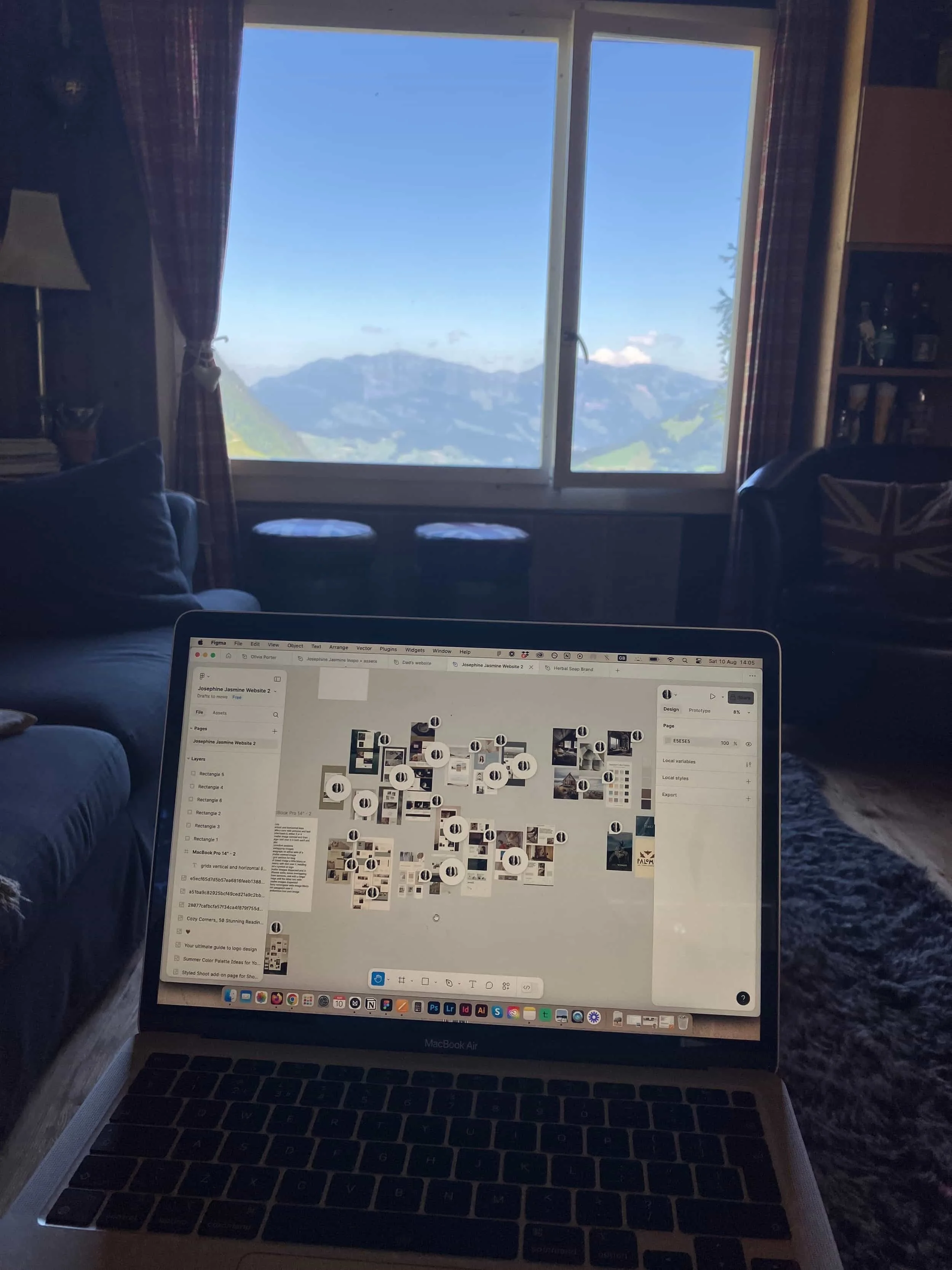 A laptop on a table displaying a design website, with a large window showing mountains and a blue sky in the background.