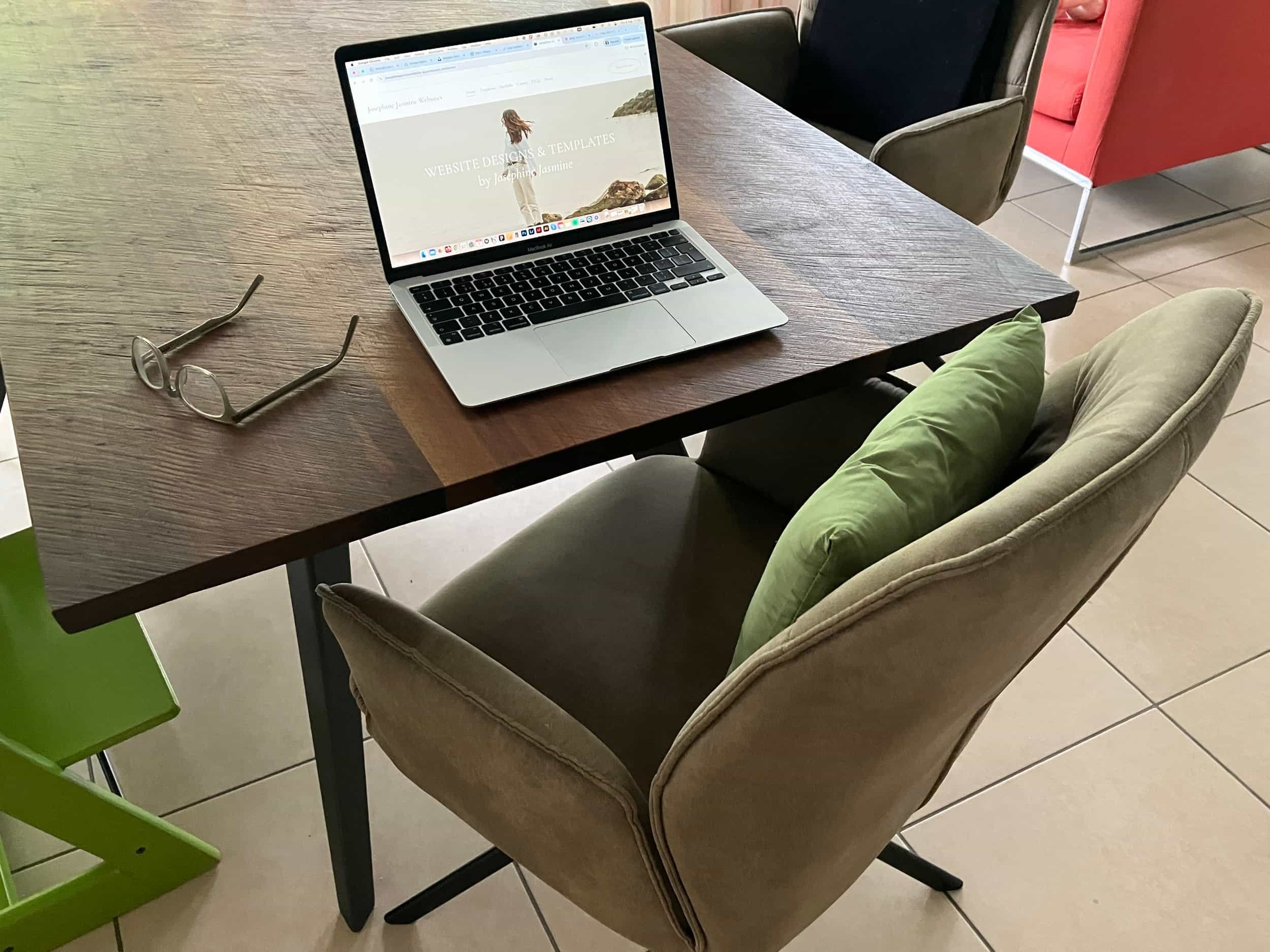 Wooden table with an open laptop displaying a website, a pair of eyeglasses, and two chairs, one beige with a green pillow and one pink, in a casual indoor setting.