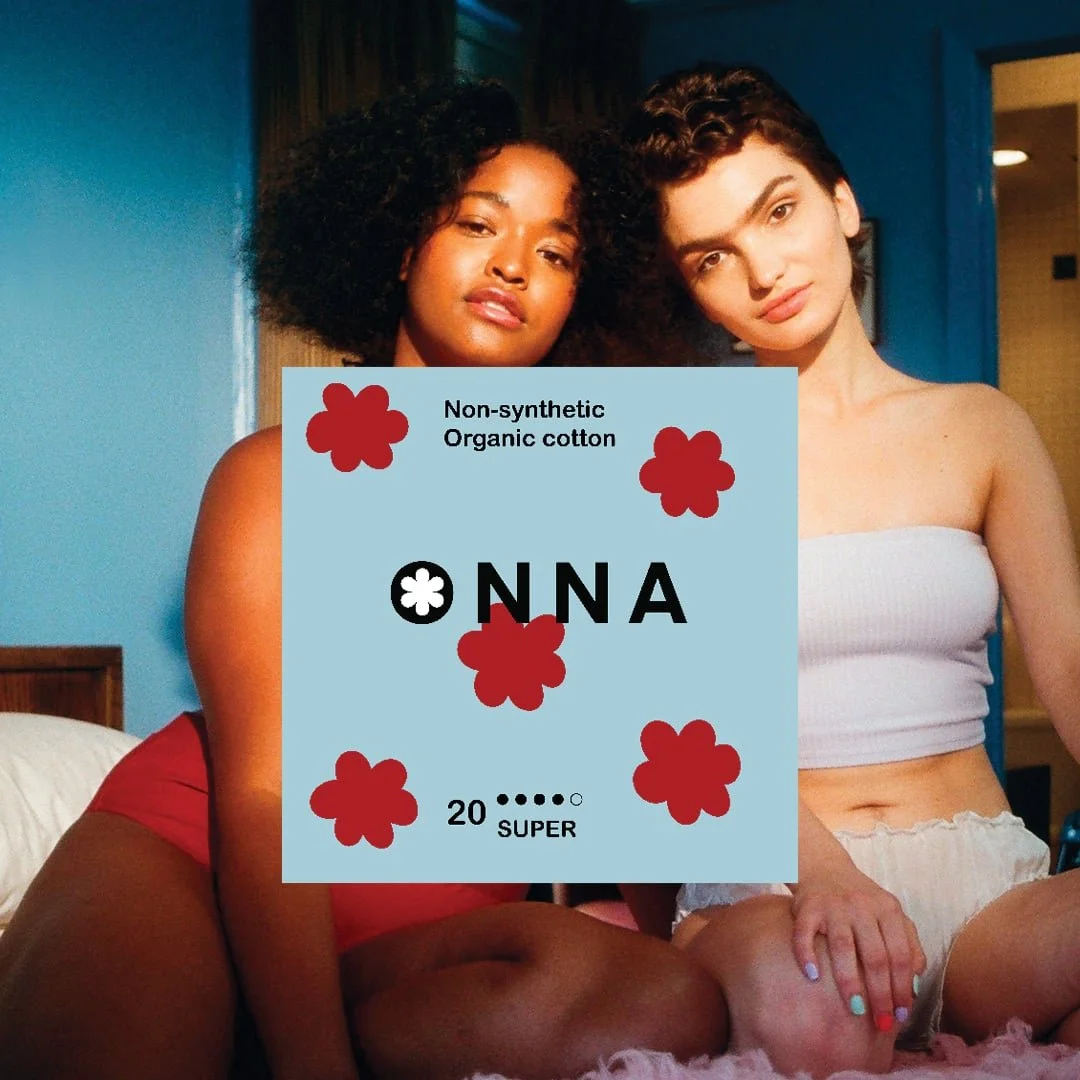Two young women sitting close together on a bed with blue walls in the background. One has curly dark hair and wears a red top, the other has wavy light brown hair and wears a white tube top. Overlaid on the image is a graphic design of a card with the word 'NONNA' and decorative red flowers.