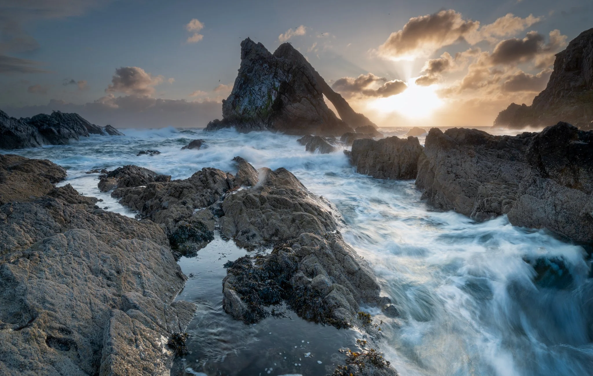 Sunset over rocky seashore with large rock formations and waves crashing against the rocks.