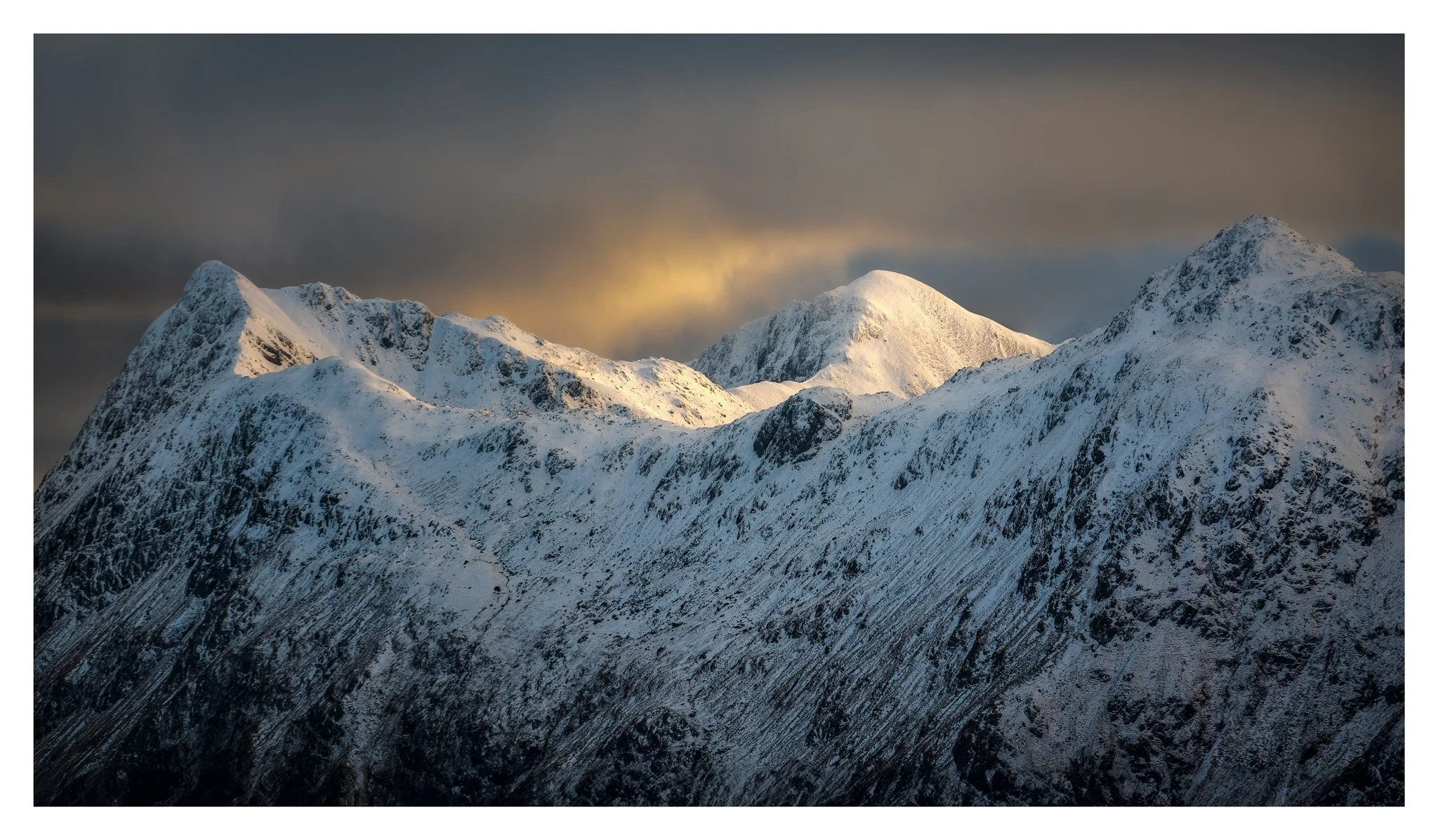 Snow-covered mountain peaks under a cloudy sky with a break in clouds showing sunlight.