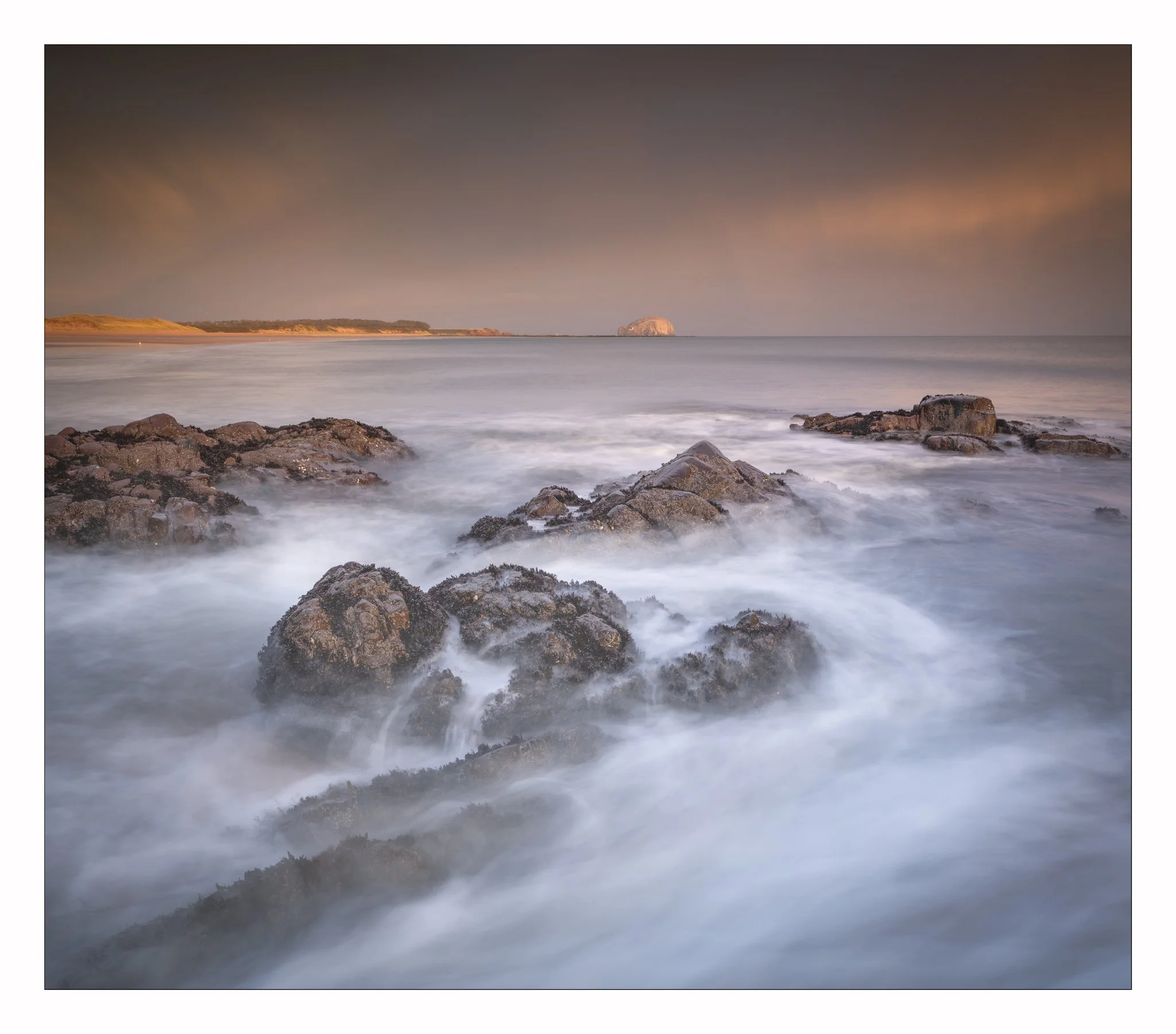 Long exposure photo of rocky shoreline at sunset with smooth ocean waves and cloudy sky in muted tones.