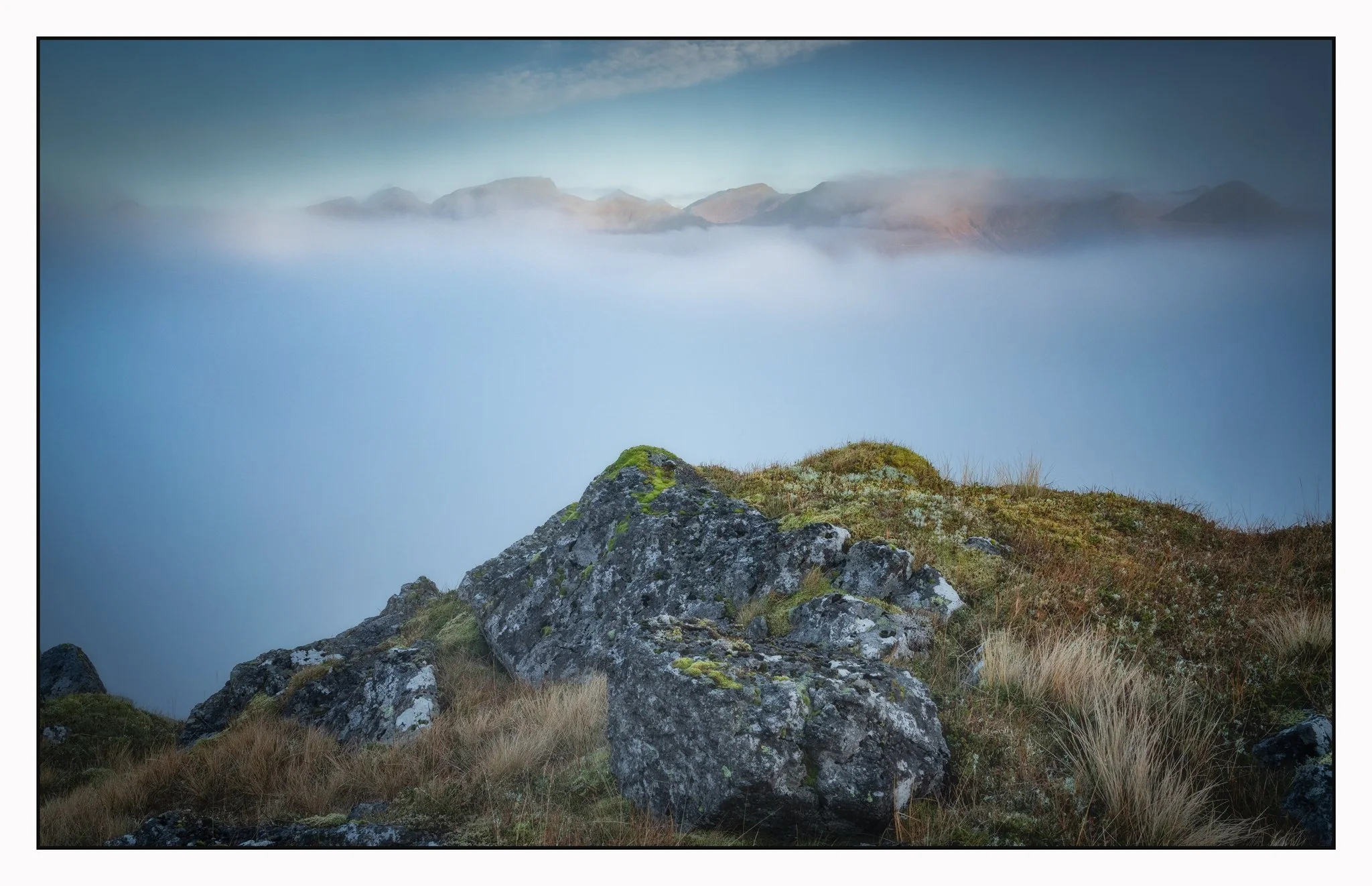 A rocky hillside with moss and grass in the foreground, fog covering a body of water, and distant mountains in the background under a partly cloudy sky.