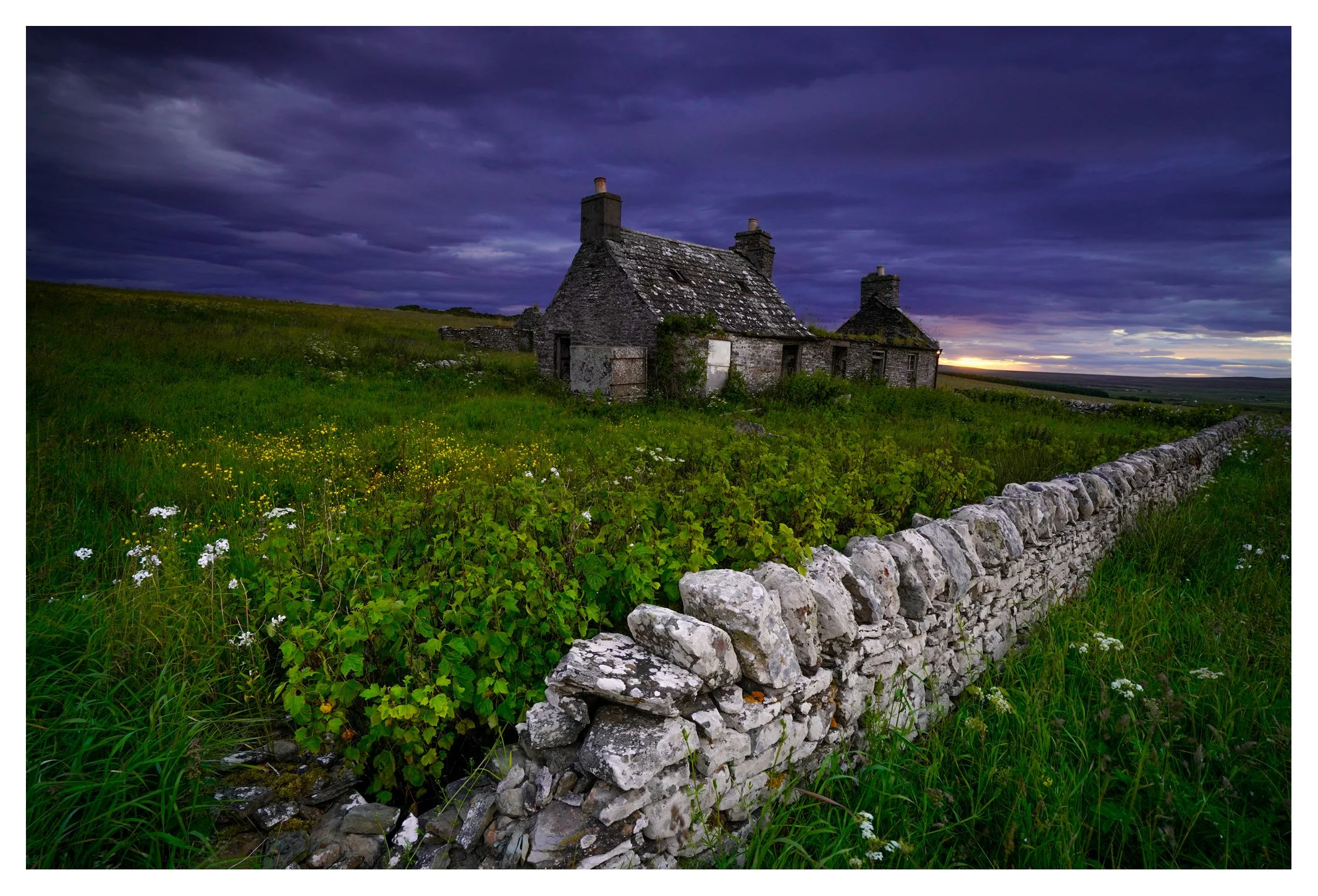 An abandoned stone cottage behind a stone wall in a grassy field with wildflowers, under a dark, cloudy sky at sunset.