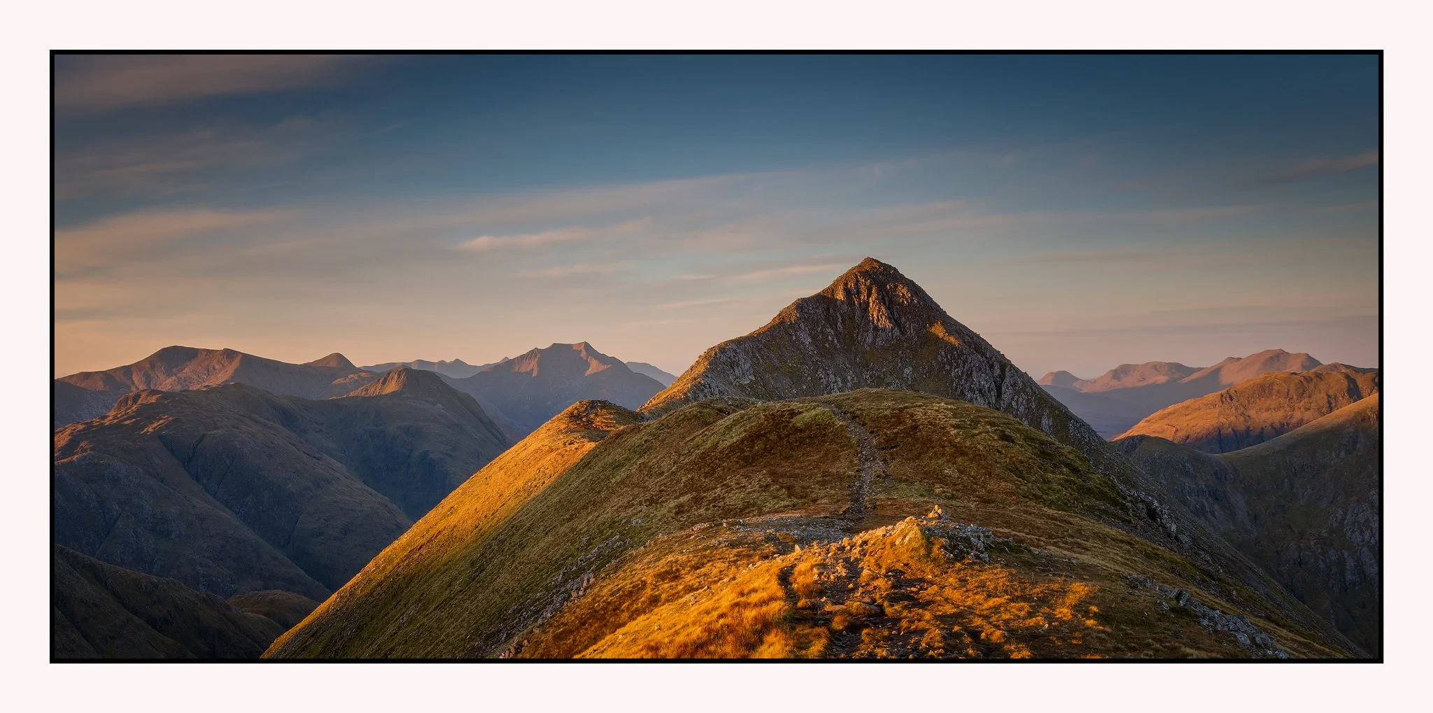 A mountain range at sunset with a prominent peak in the foreground and a winding trail leading up the hill.