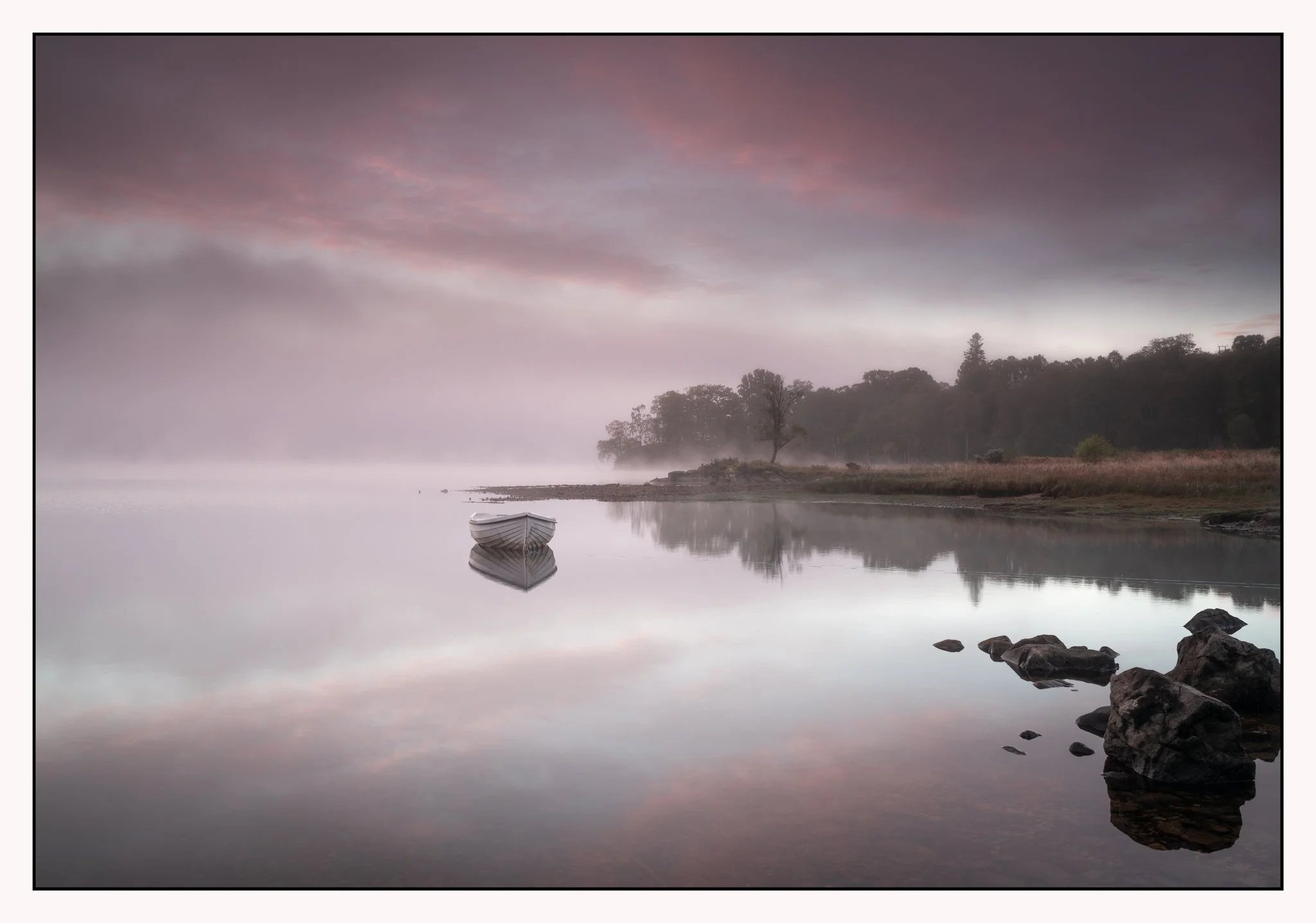 Calm lake with a small white boat reflected in the water, surrounded by rocks, with a foggy shoreline and trees under a cloudy sky during dawn or dusk.