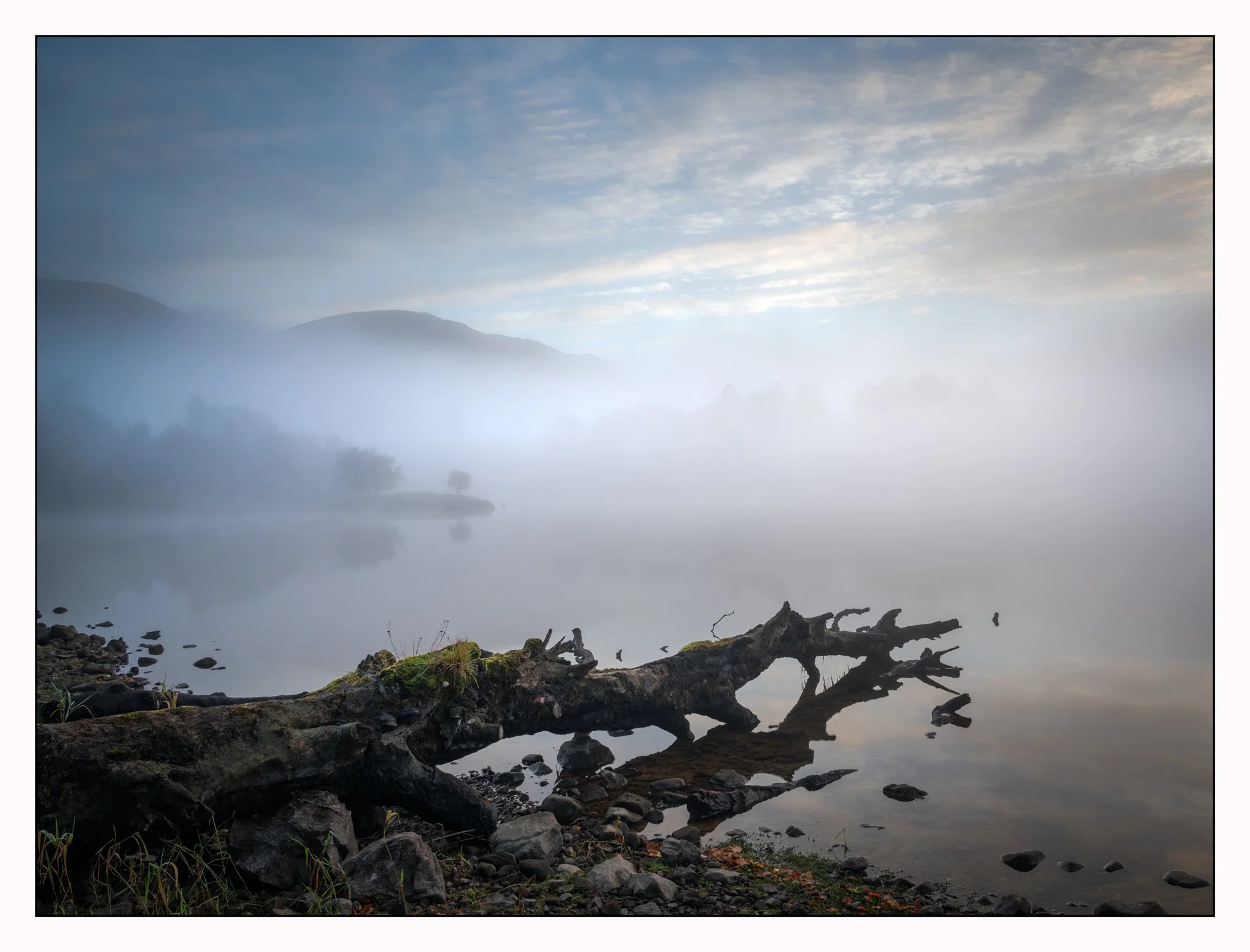 A foggy lakeshore with calm water, rocky shoreline, fallen tree, and distant mist-covered hills under a cloudy sky.