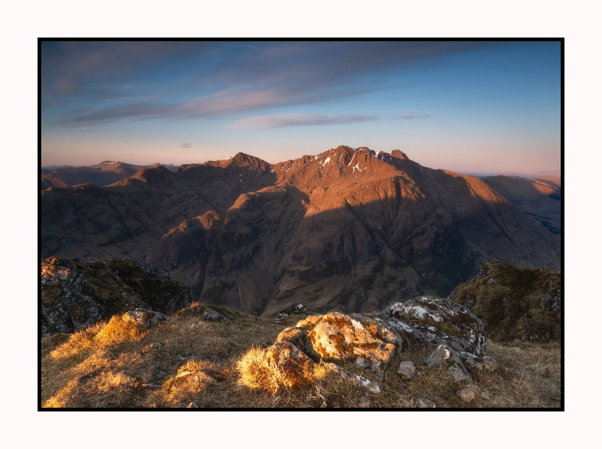 Sunset over rugged mountain range with snow patches, rocky foreground, and blue sky with light clouds.