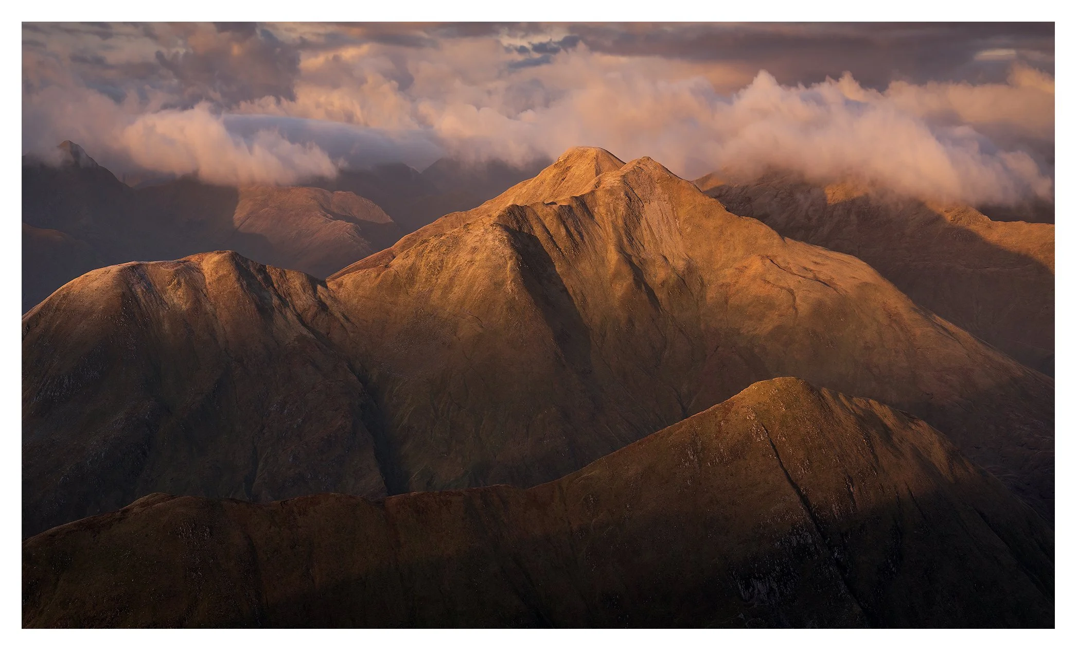 Sunlit mountain range with rugged peaks and clouds in the sky.