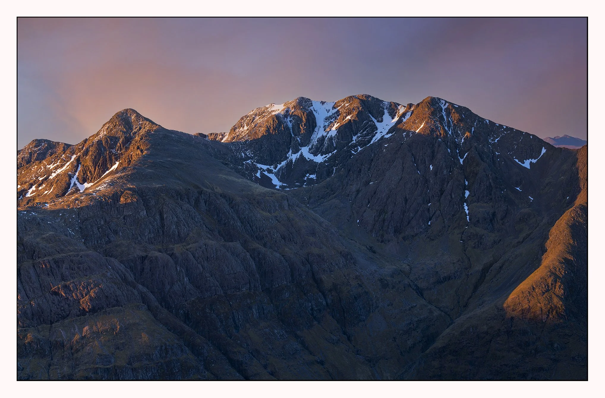 Mountain range during sunset with rocky peaks and patches of snow.