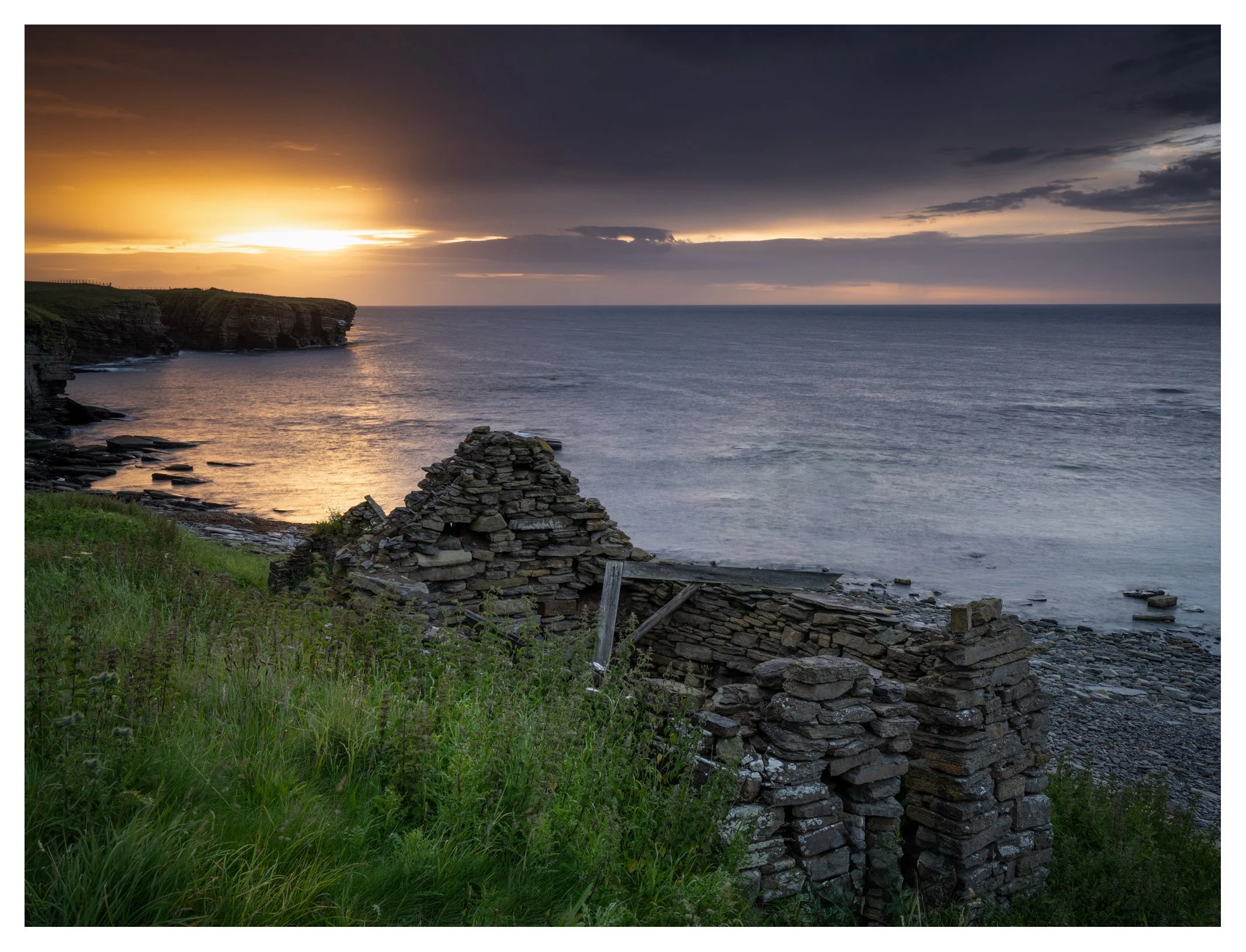 Sunset over the ocean with rocky cliffs and a grassy hillside in the foreground, and an old stone structure partially collapsed.