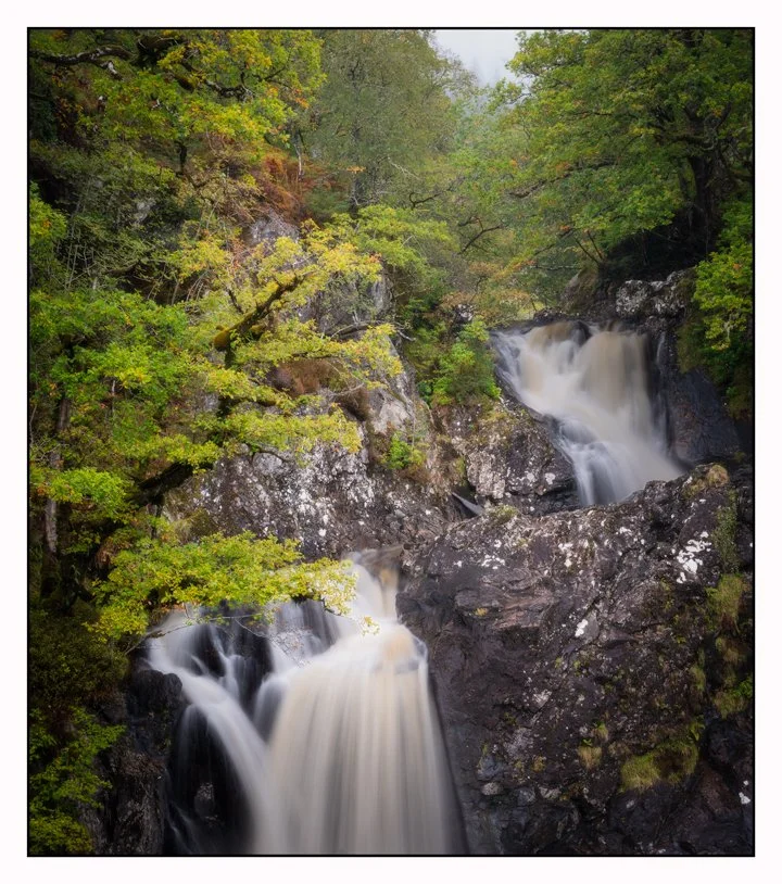 A multi-tiered waterfall flowing over dark rocks surrounded by lush green trees and foliage.