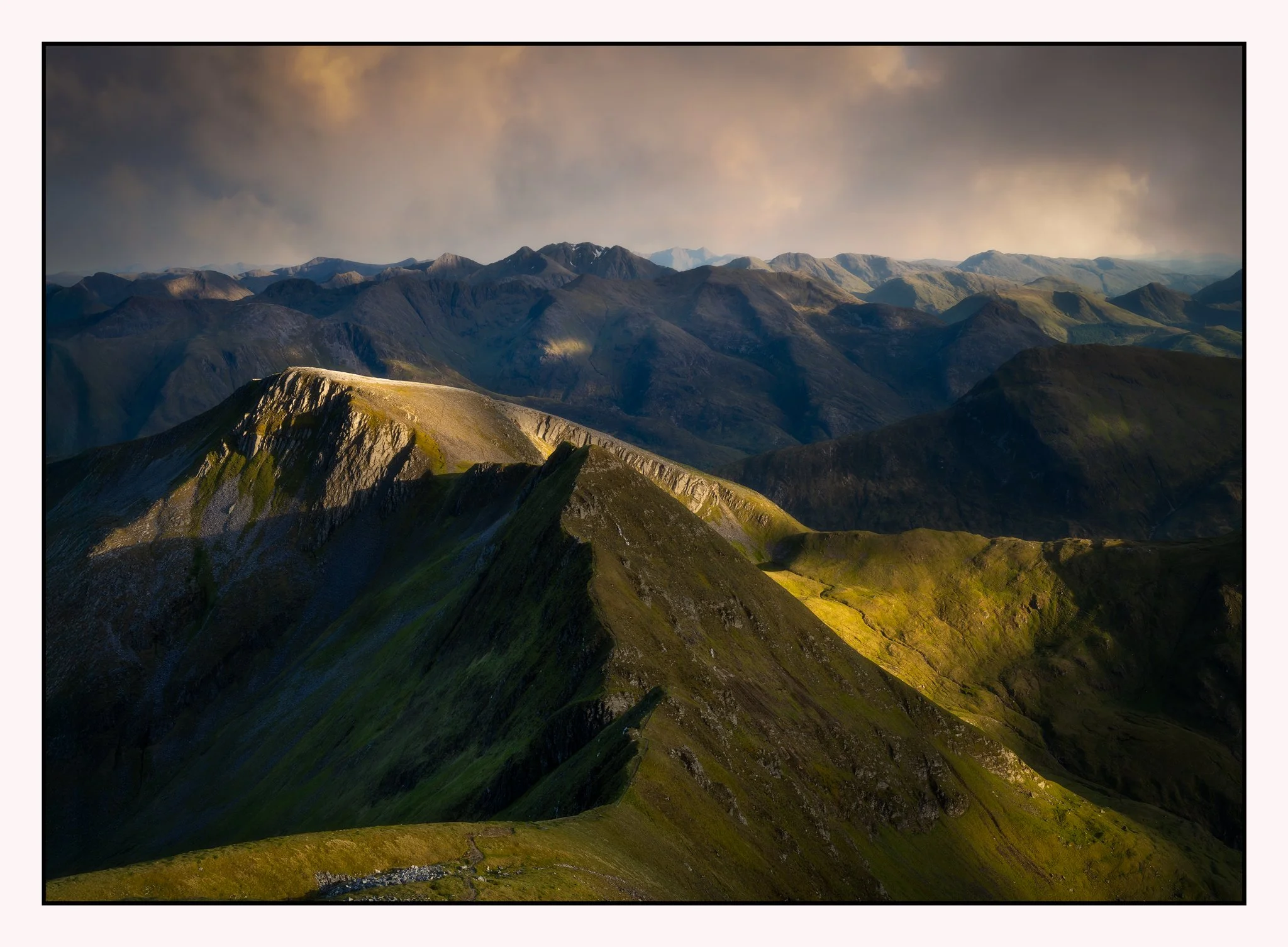 Sunlit mountain ridges with green slopes and deep valleys under a cloudy sky.