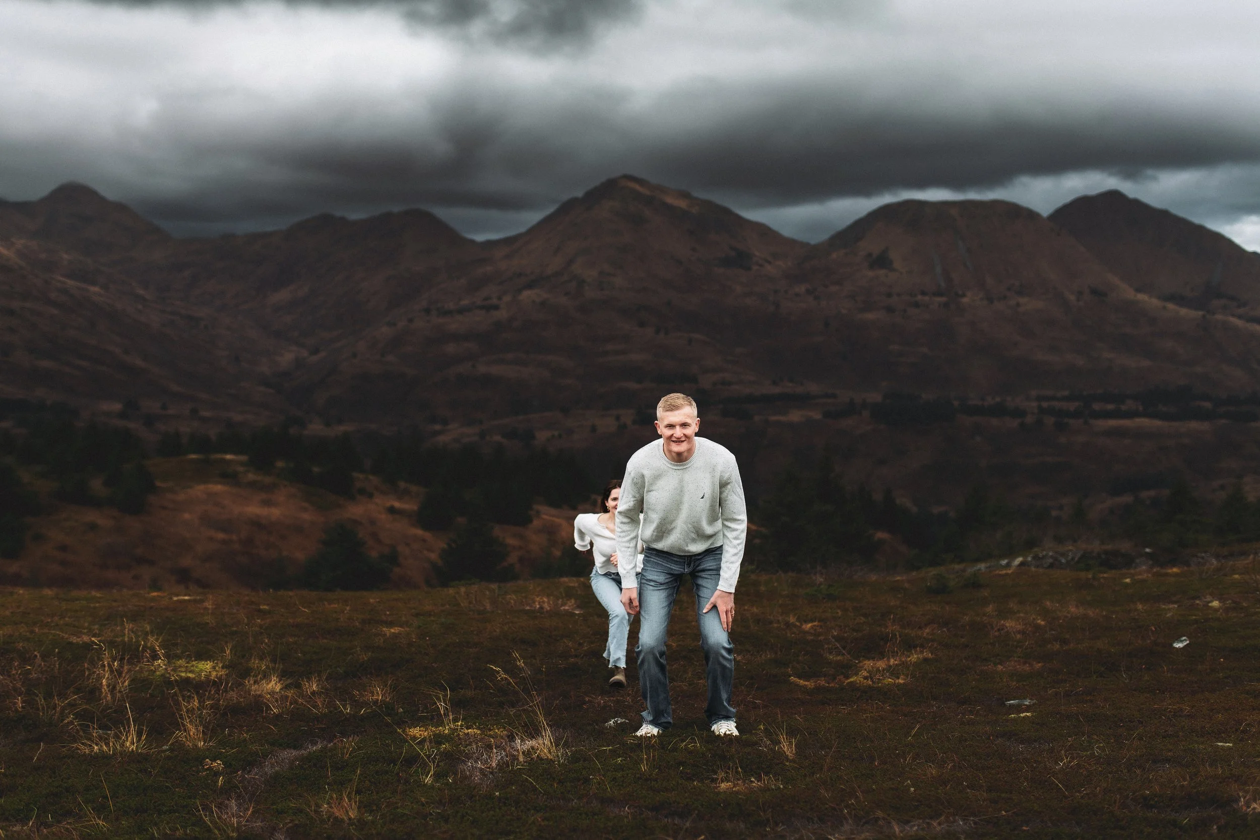Two people, a man in the foreground and a woman behind him, are running or playing on a grassy field with mountainous terrain and dark cloudy sky in the background.