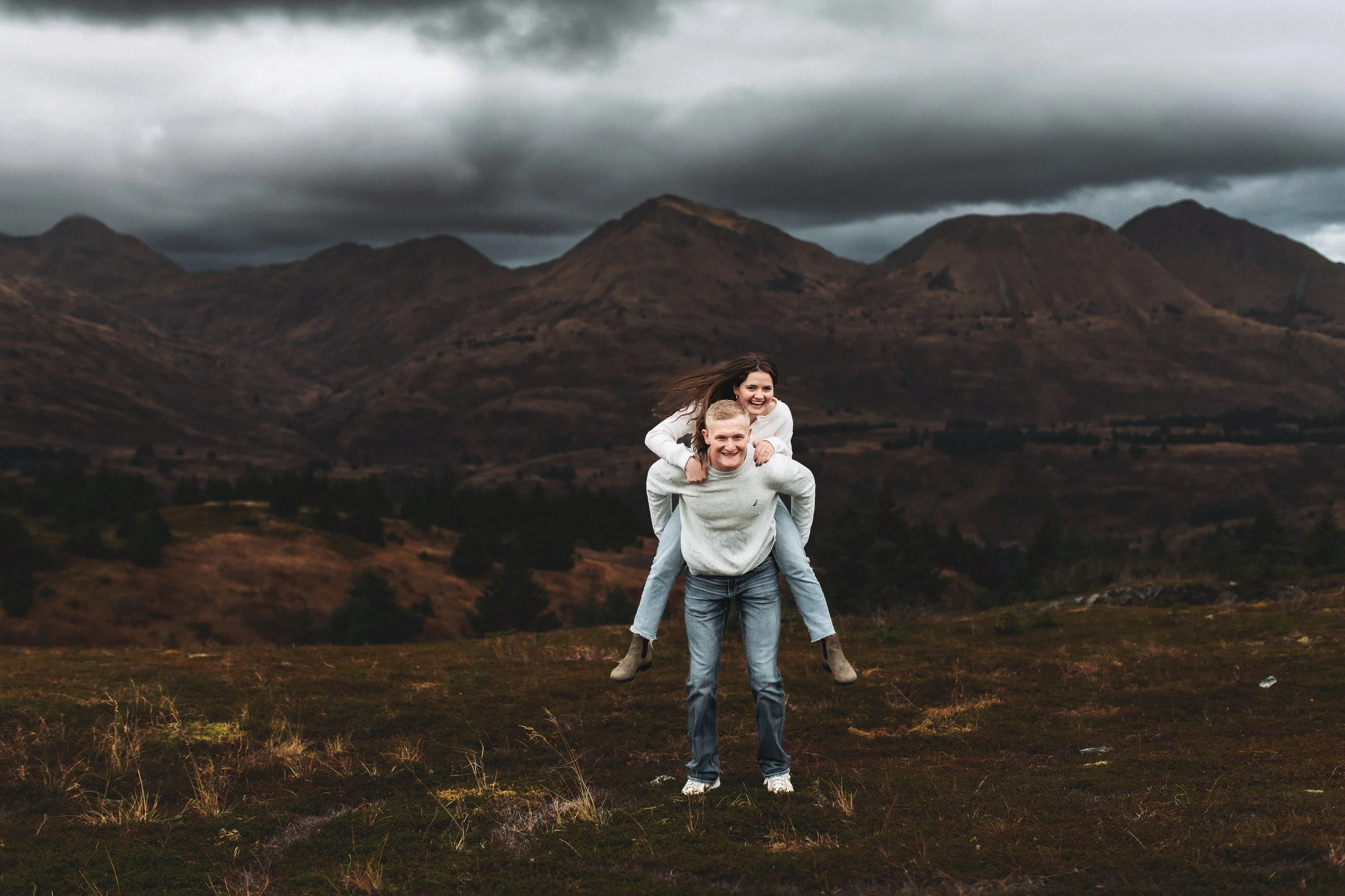 A man giving a woman a piggyback ride in a mountainous outdoor area with dark clouds overhead.