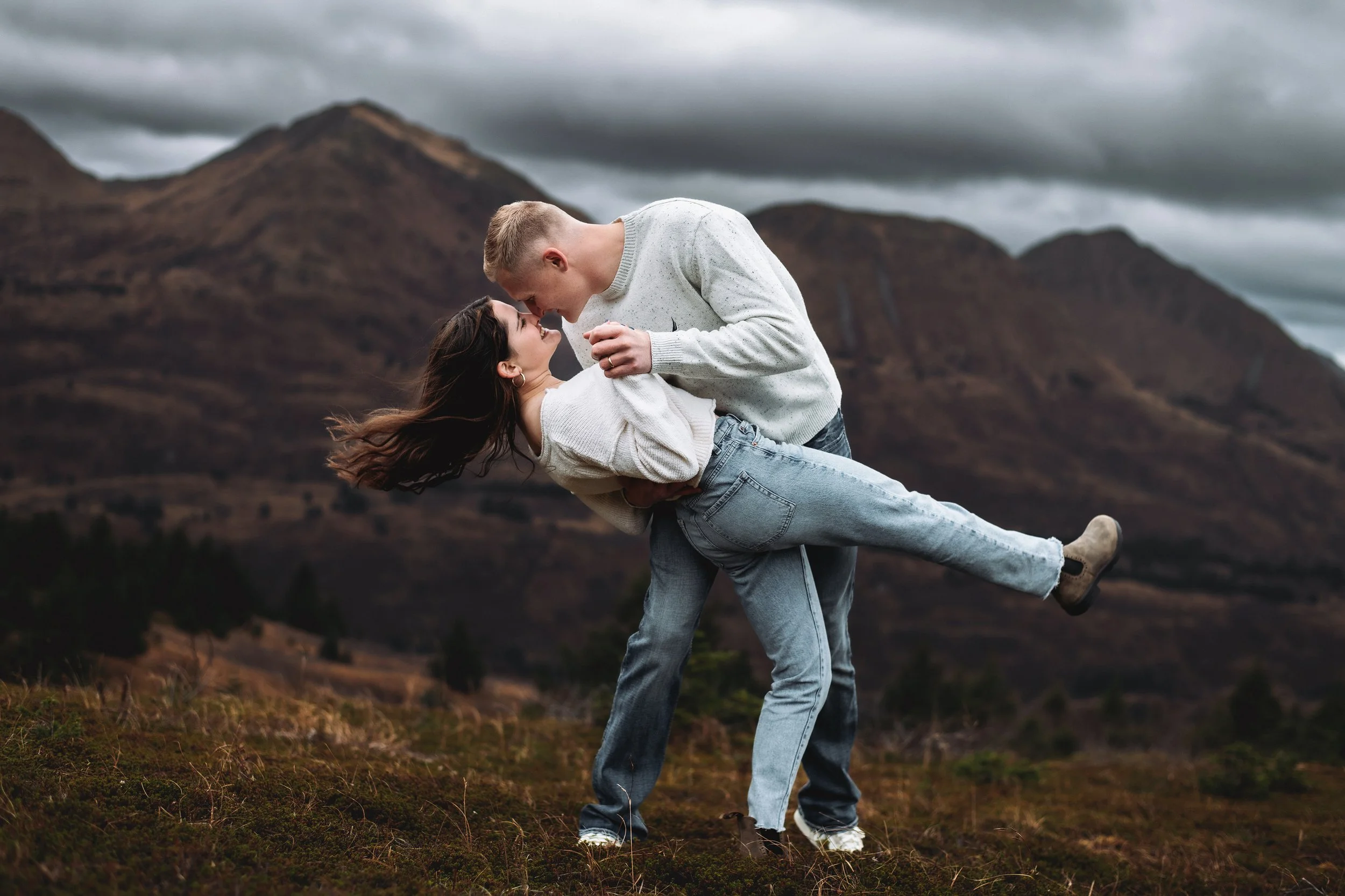 A couple on a mountain landscape, with the man holding the woman as she leans back, both looking at each other.