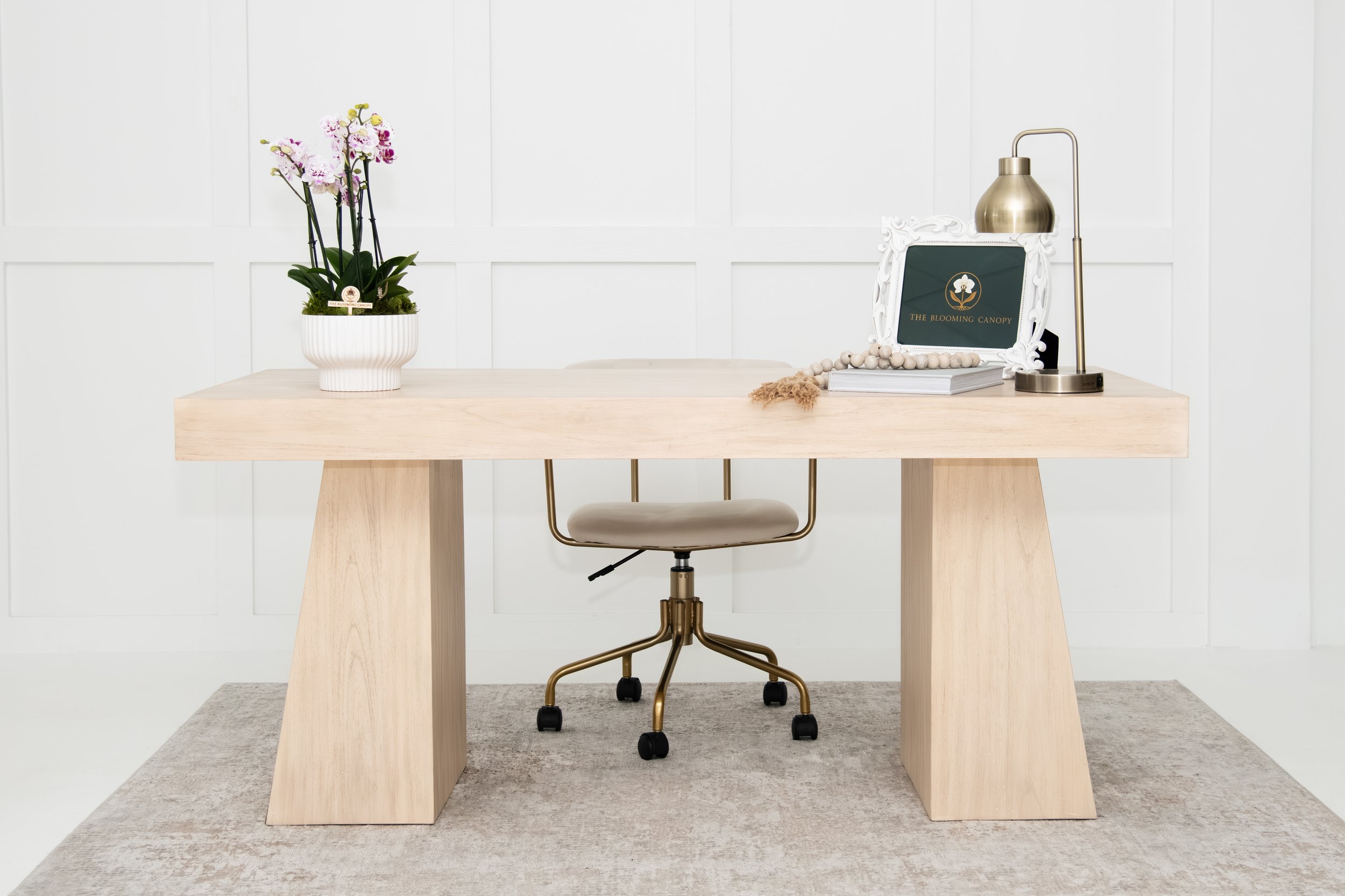 Minimalist office desk with a potted orchid, a framed sign reading 'The Blooming Canopy,' a stack of books, a decorative bead strand, and a brass desk lamp, set against a white paneled wall.