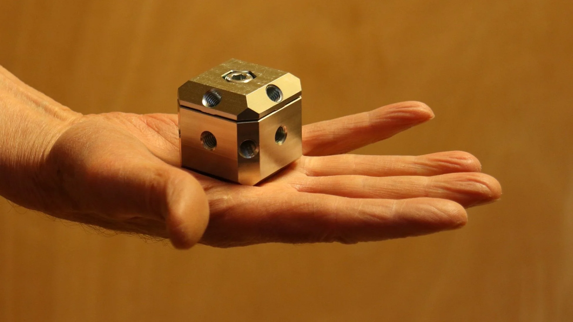 A human hand holding a metallic cube with multiple threaded holes on each face, against a blurred brown background.