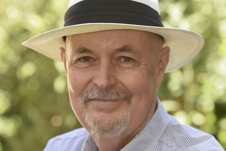 Close-up of a smiling man wearing a wide-brimmed hat and striped shirt outdoors with green foliage in the background.