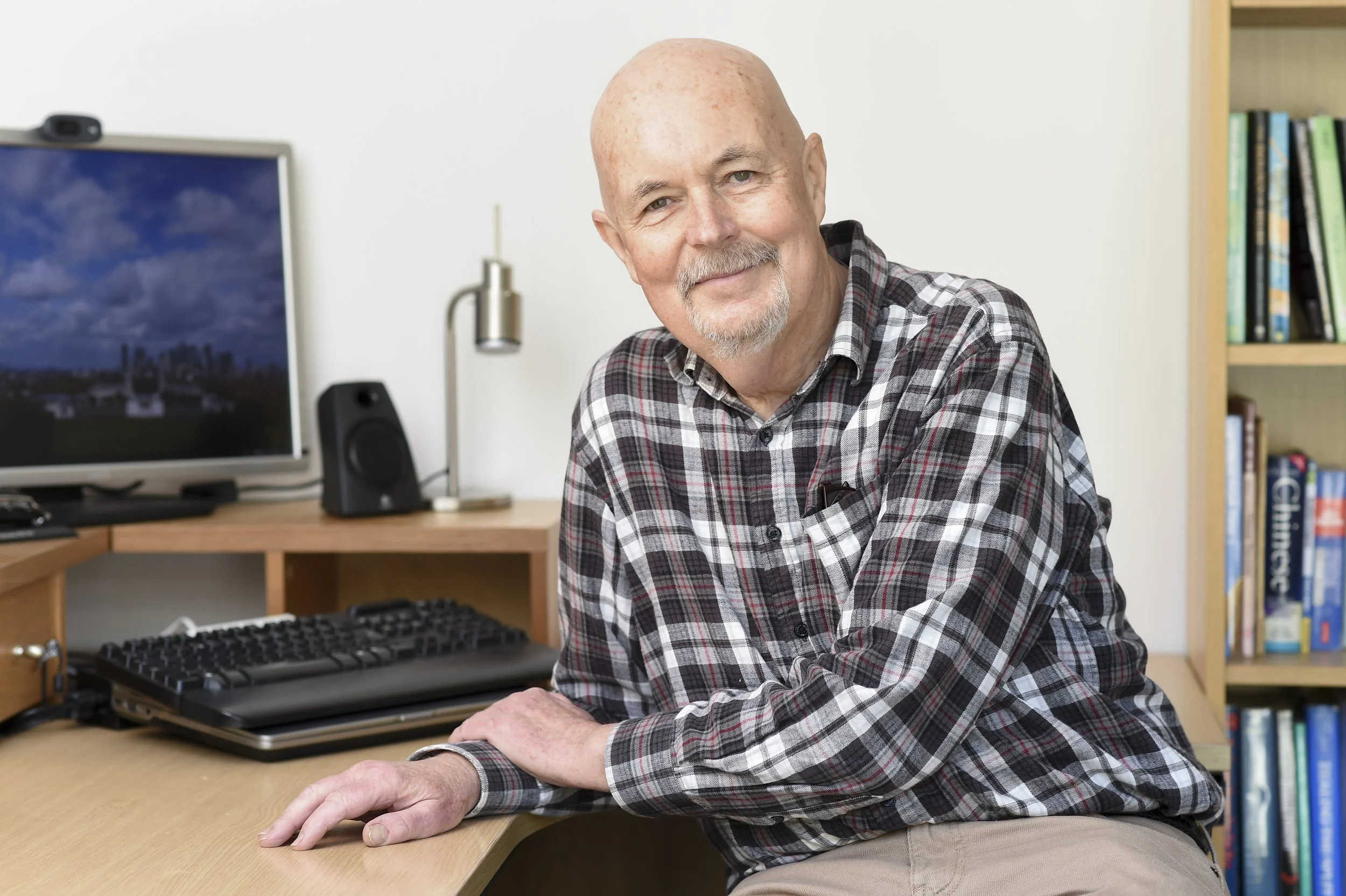 A smiling elderly man with a shaved head, gray goatee, and light skin, wearing a gray and black checkered shirt, sitting at a wooden desk in a room with a bookshelf and a computer monitor in the background.