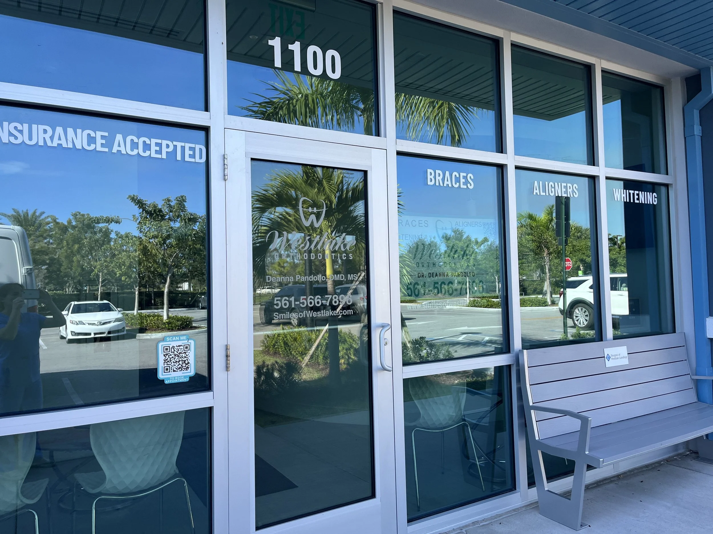 Front window of a dental orthodontics office with blue-tinted glass, displaying signs for insurance acceptance, braces, aligners, and whitening, with a bench outside and reflections of trees, cars, and a person taking the photo.