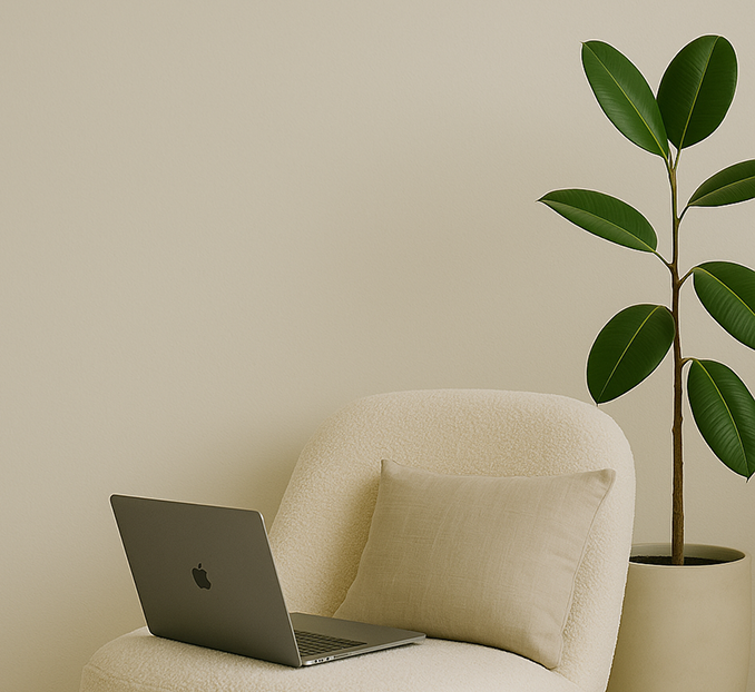 A cream-colored chair with a pillow, a laptop, and a tall potted plant with large green leaves against a plain beige wall.