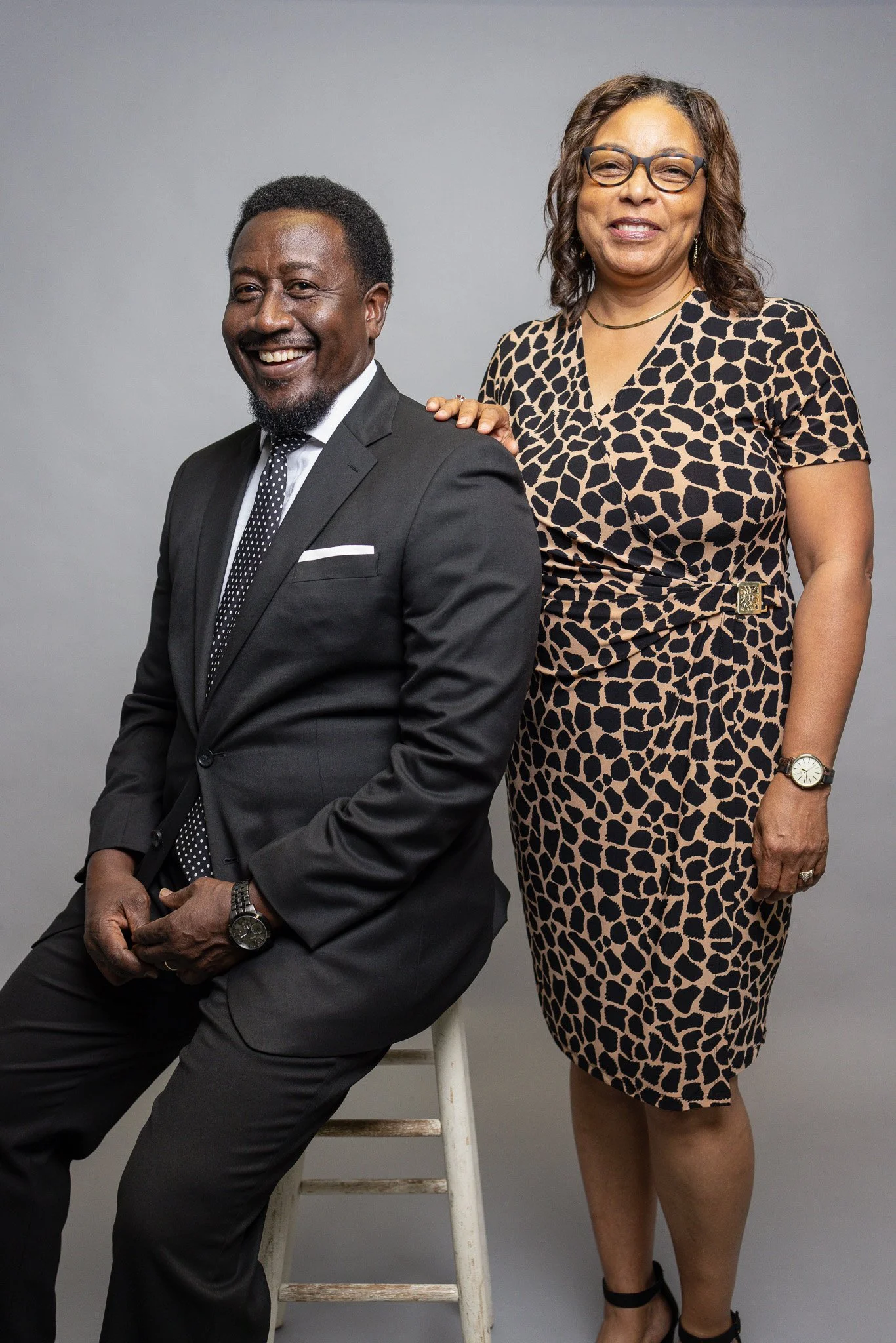 A man in a black suit sitting on a stool and a woman in a leopard print dress standing behind him, both smiling against a gray background.