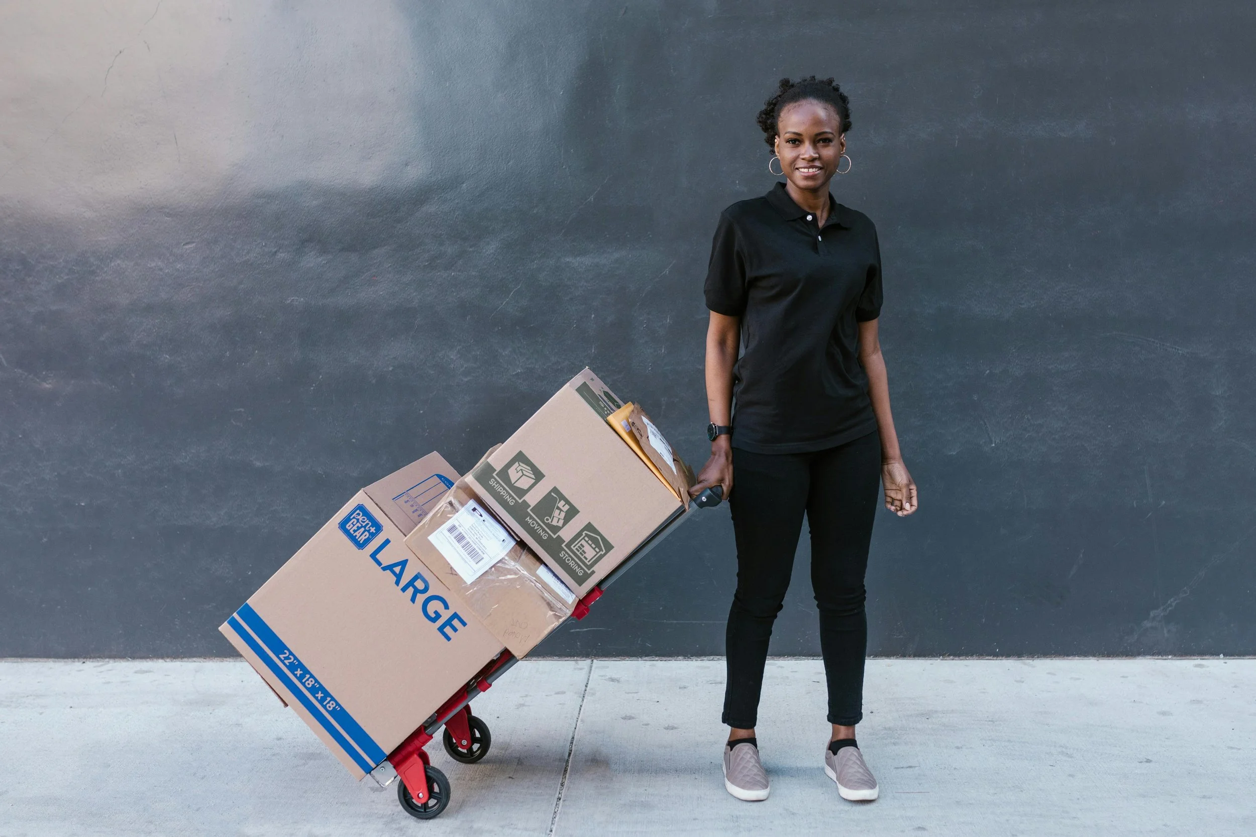 A woman in black clothing standing outside a dark gray wall, holding a red hand truck loaded with large boxes, including a 22-inch by 18-inch by 18-inch large cardboard box and smaller boxes.
