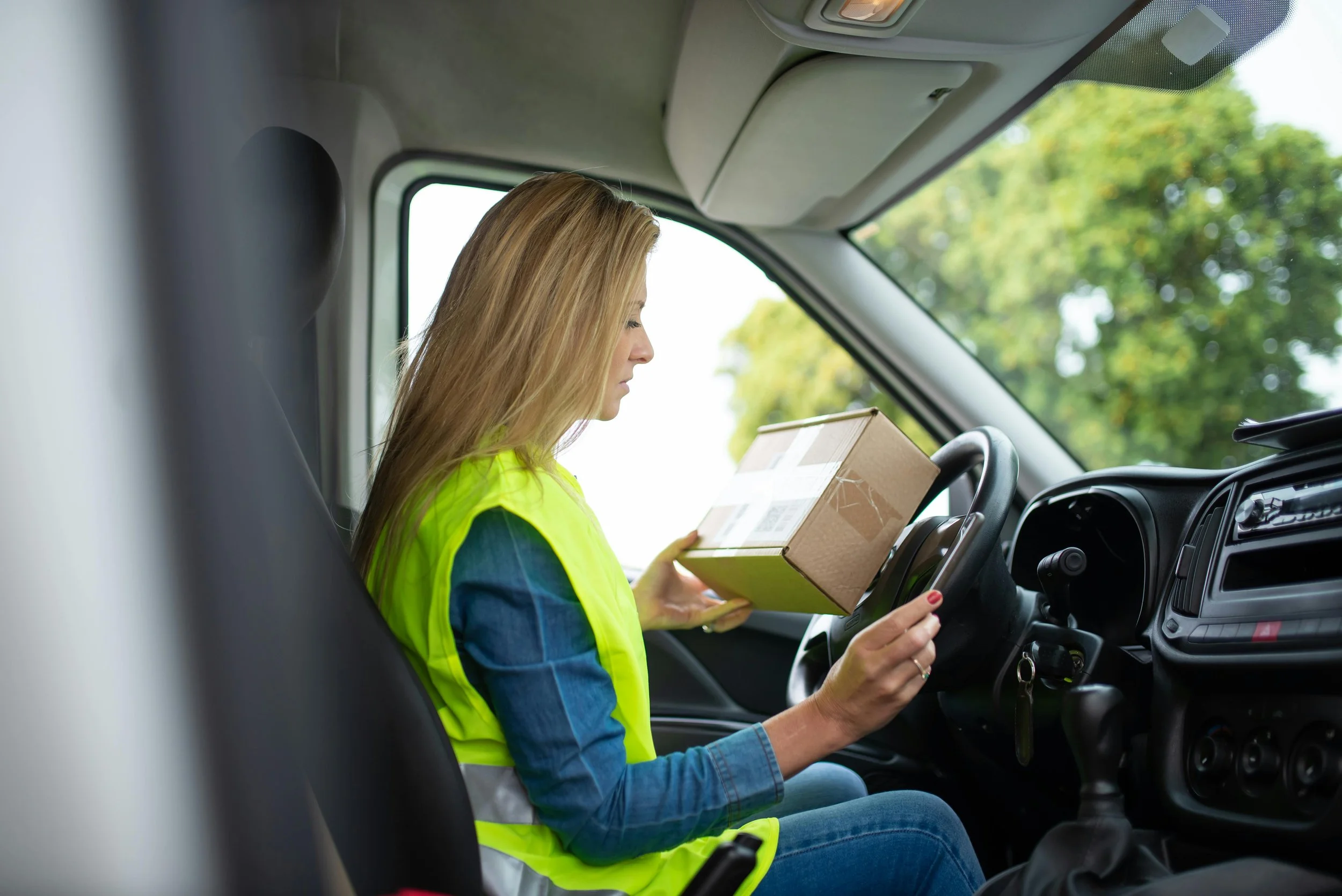 A woman with blonde hair wearing a yellow safety vest sitting in the driver's seat of a truck, holding a cardboard box, with trees visible outside the windshield.