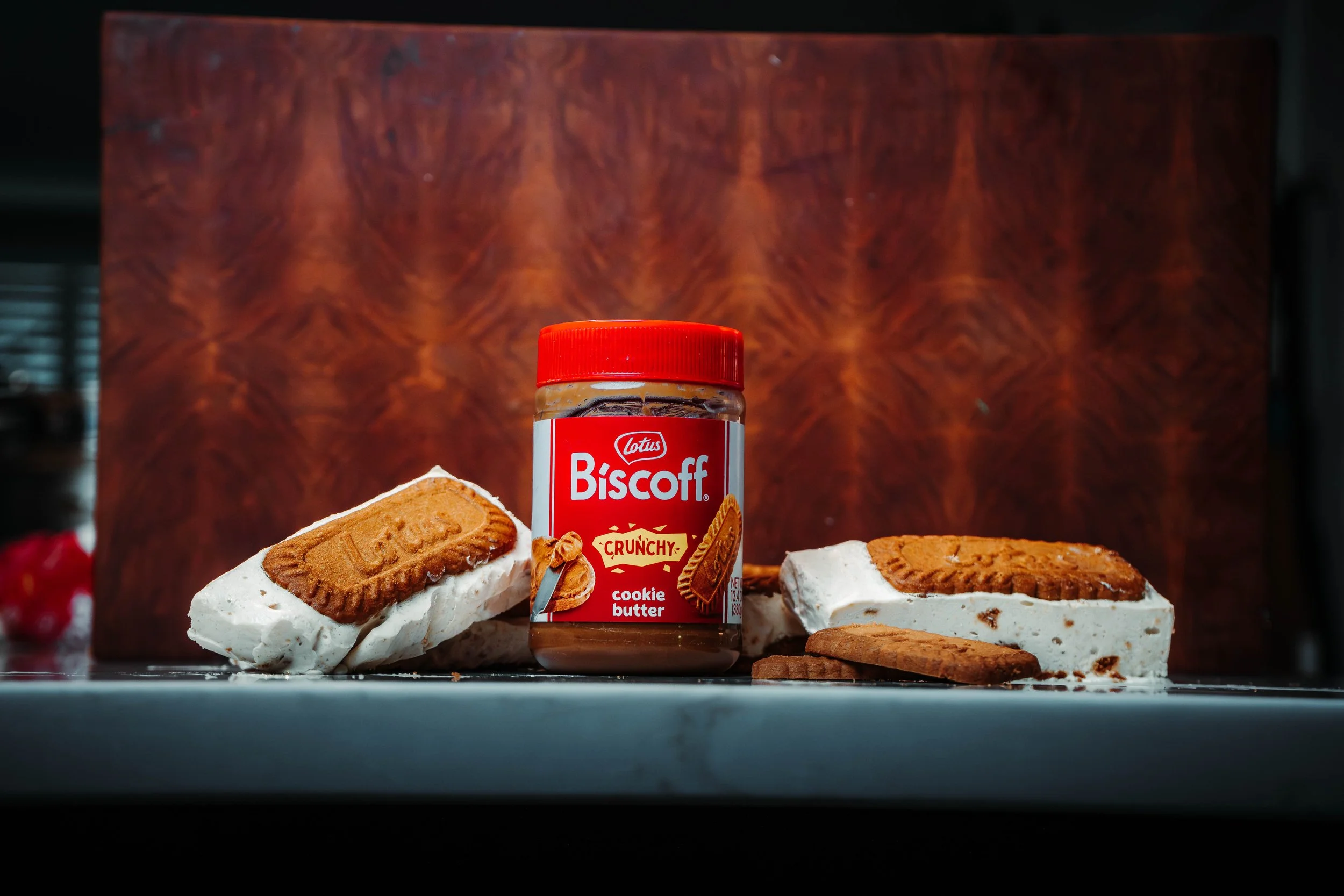 Container of Lotus Biscoff crunchy cookie butter surrounded by biscuits and cream cheese with biscuits on top, on a reflective surface with a wood grain background.