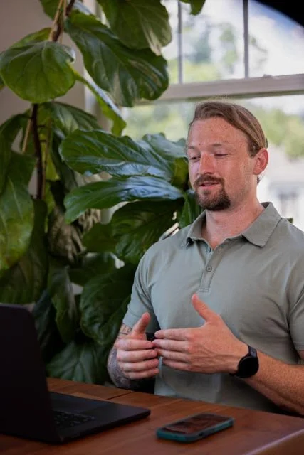 Man with a beard sitting at a table in front of a laptop, gesturing with his hands, surrounded by large green plants near a window.