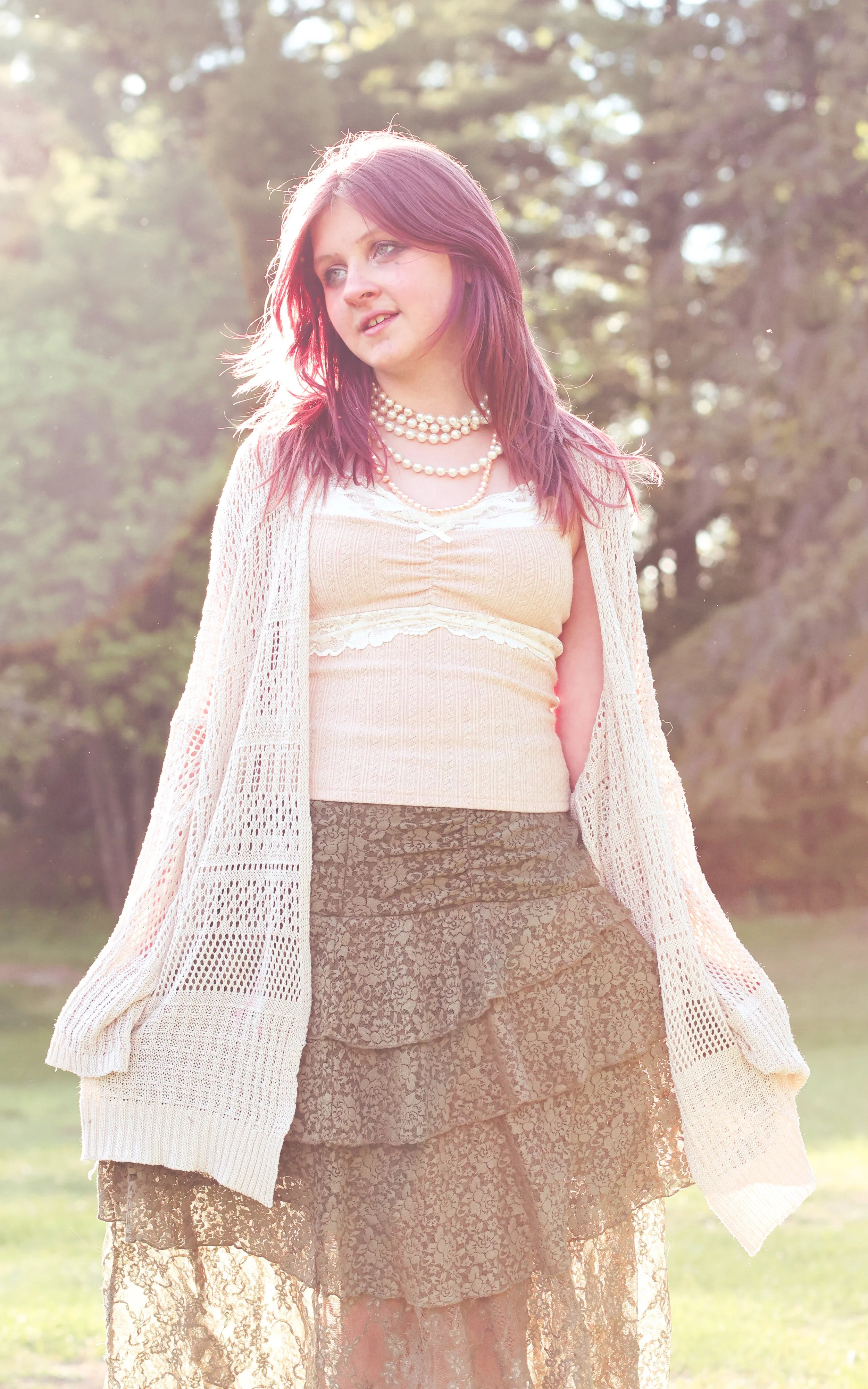 Young woman with shoulder-length purple hair wearing layered pearl necklaces, a pastel yellow top, a white open-knit cardigan, and a lace skirt standing outdoors in sunlight with trees in the background.