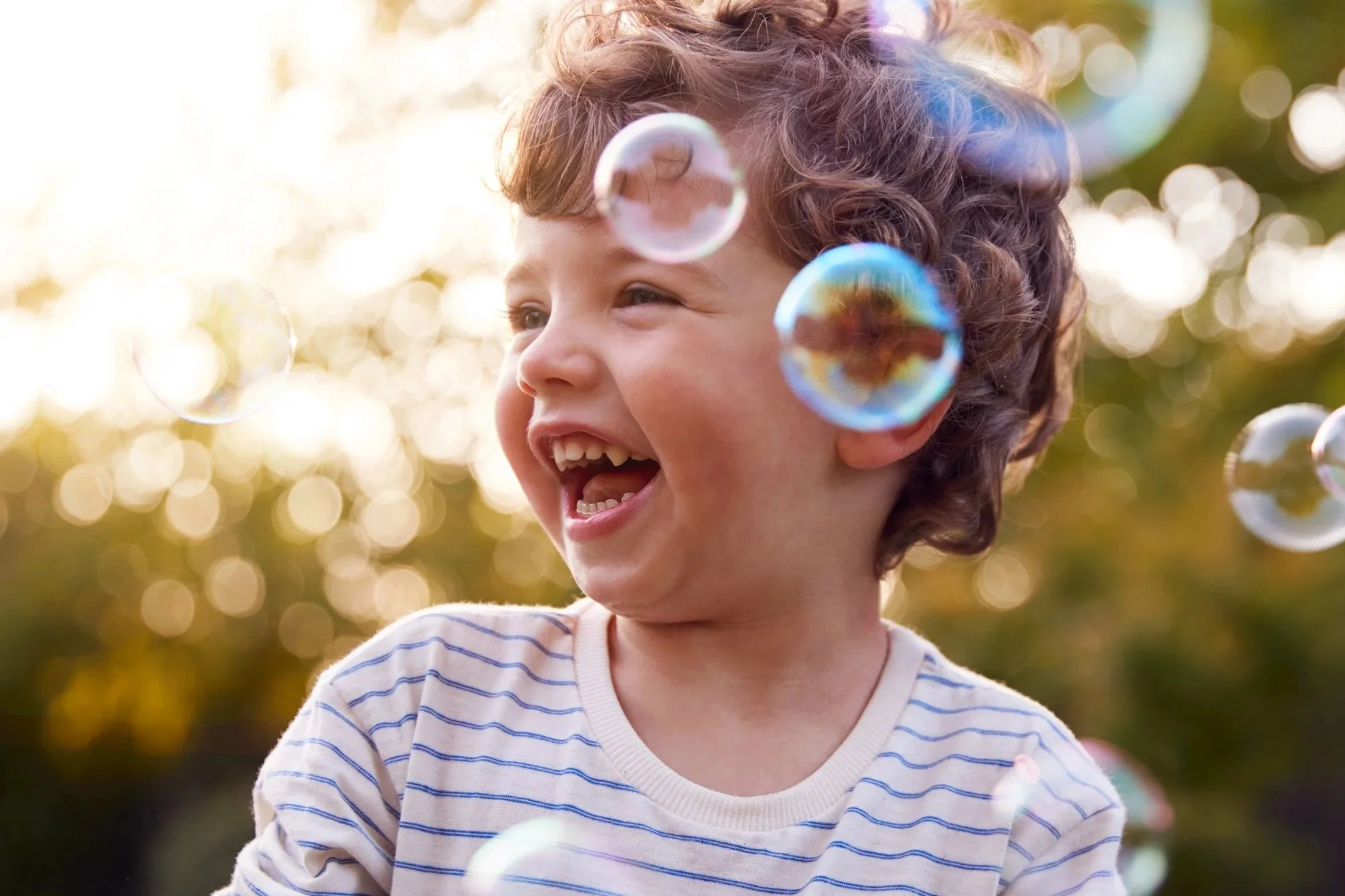 A young boy with curly hair smiling and looking to the side outdoors, surrounded by floating bubbles with a blurred background of sunlight and trees.