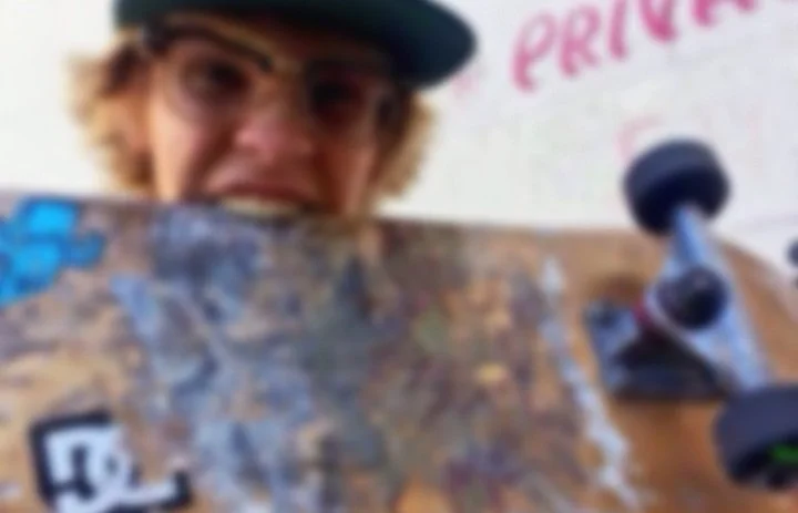 Blurry image of a person with glasses and a cap biting a skateboard.