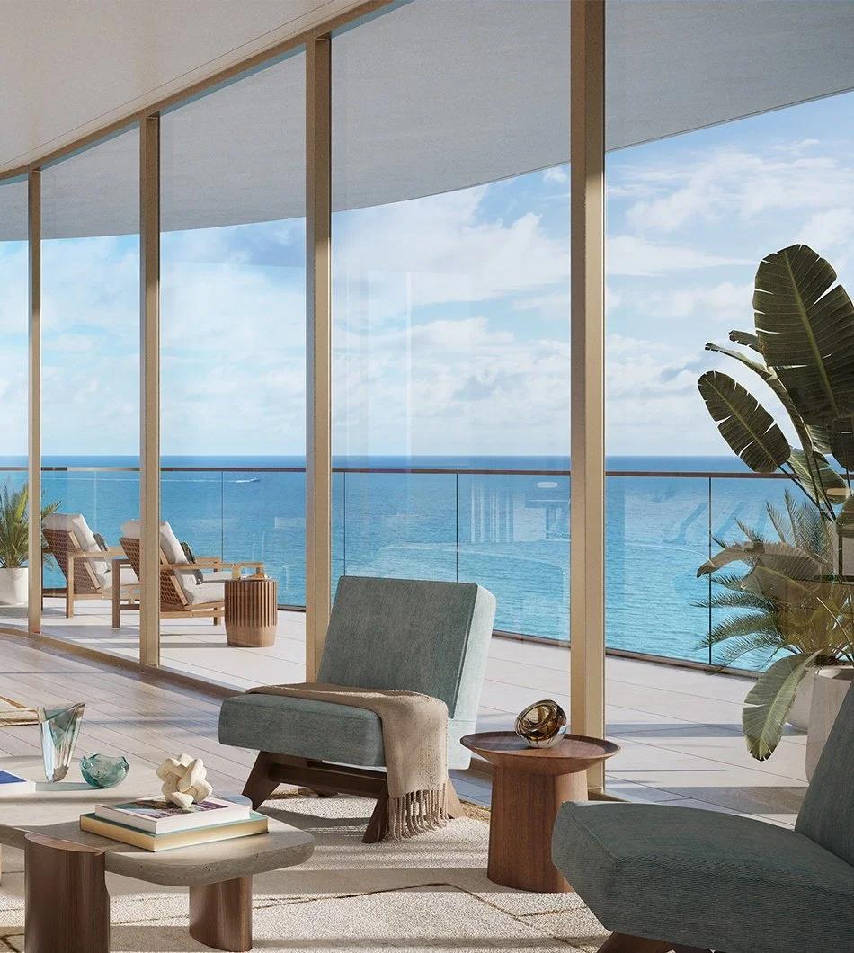 Living room with large glass windows overlooking the ocean, furnished with light blue armchairs, a wooden side table with decorative objects, and a potted plant, with a balcony and ocean view in the background.