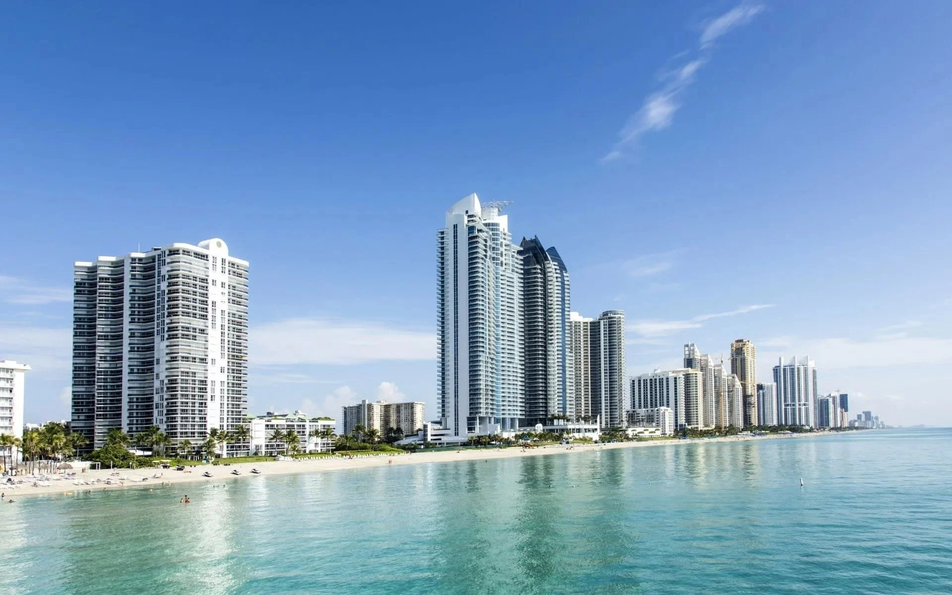 Skyline view of tall modern skyscrapers along a beach with clear turquoise water and a blue sky.