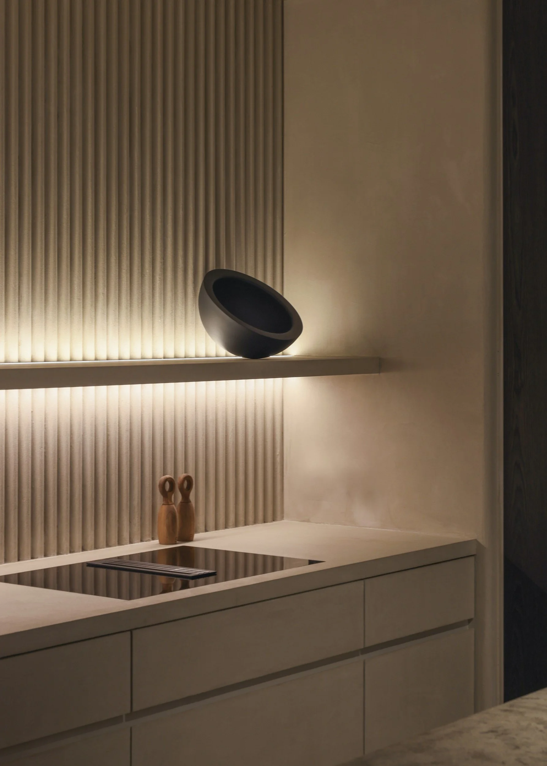 Minimalist kitchen with beige cabinetry and a beige countertop, featuring two small wooden vases on the counter, a black angled speaker on a shelf above, backlit textured wall paneling, and a dark door to the right.