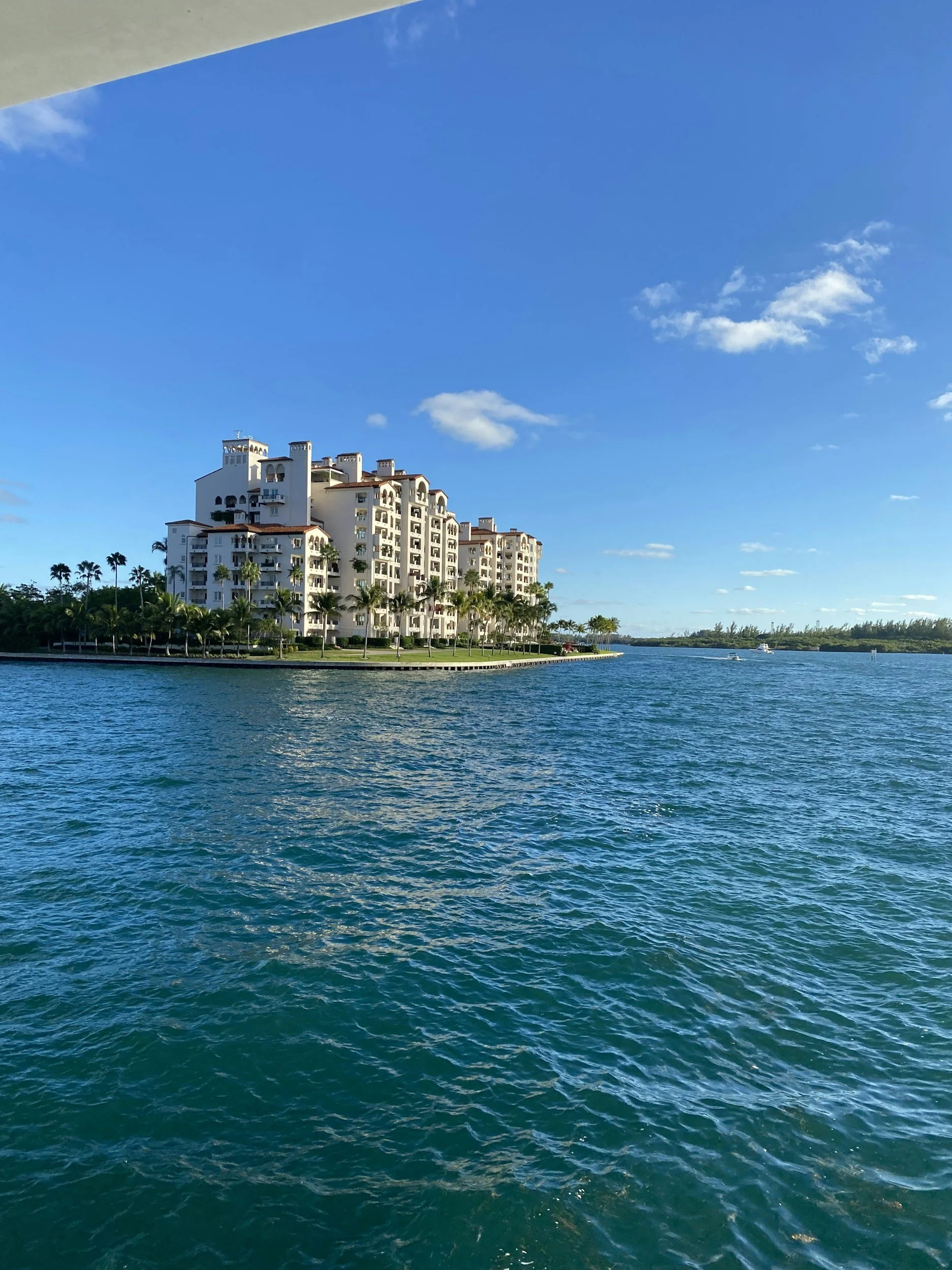 A white multi-story building by a water body, with palm trees lining the shore, under a blue sky with a few clouds.