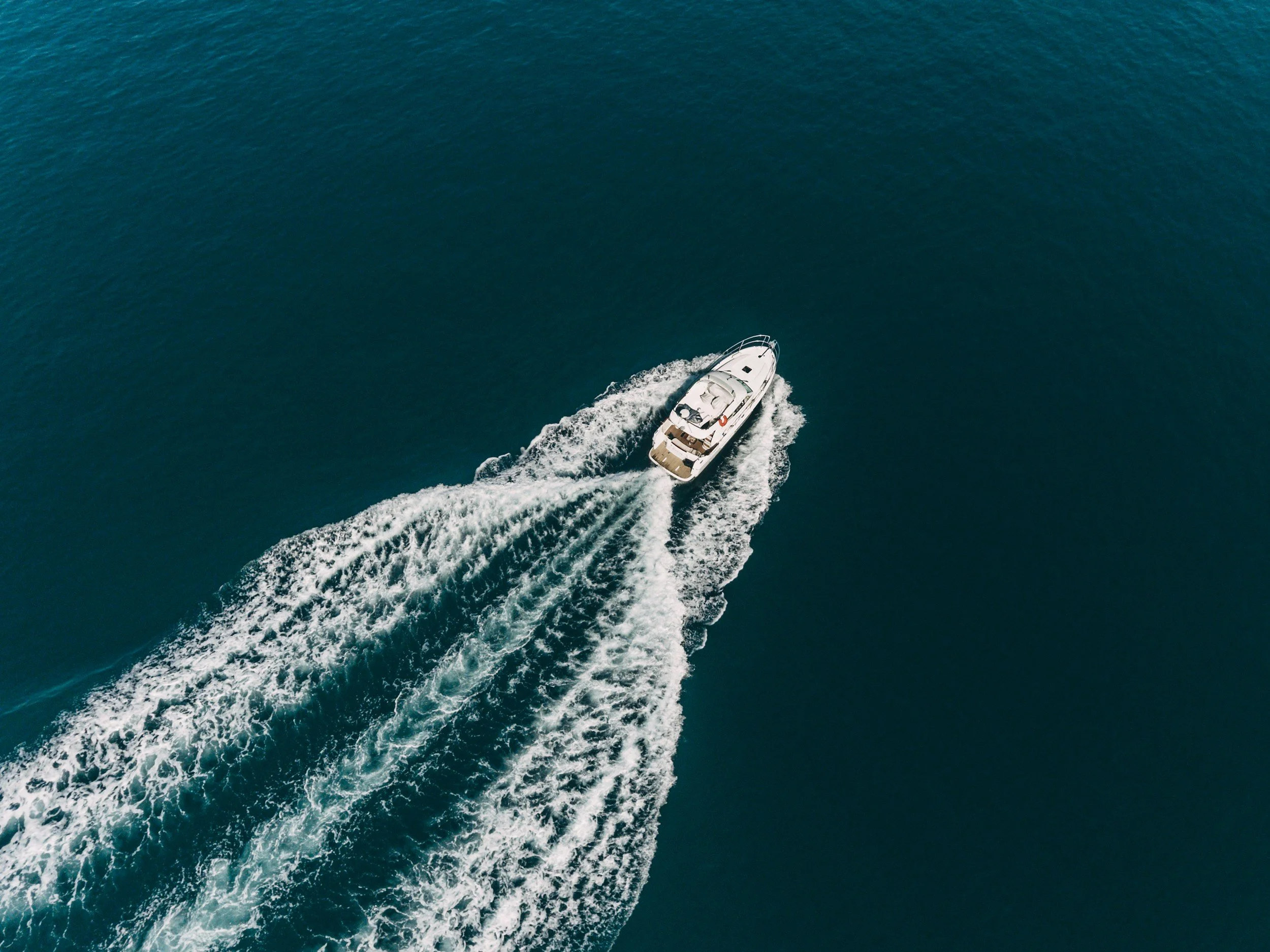Aerial view of a white yacht moving through deep blue ocean, creating waves behind it.