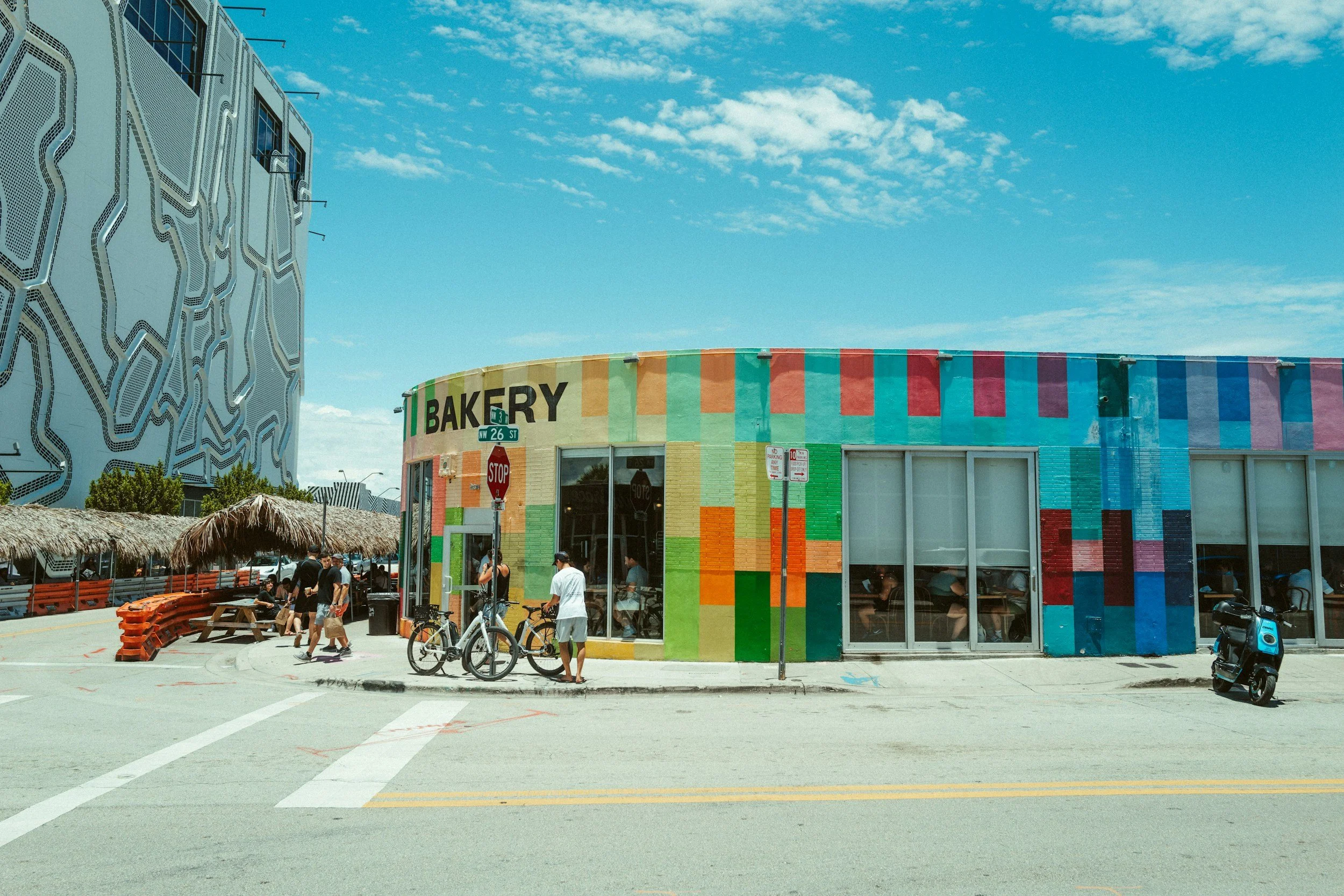 Colorful bakery building on a corner with people, bicycles, and a scooter outside, under a blue sky with scattered clouds.