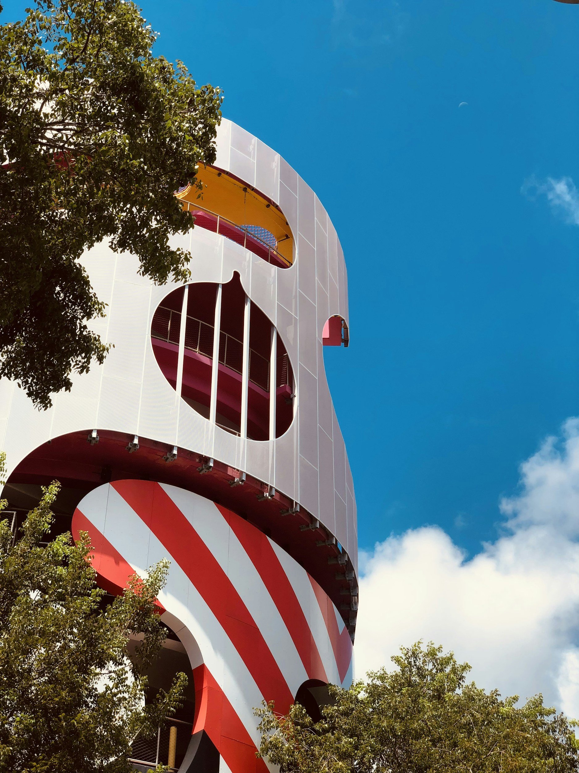 A modern, colorful building with a candy cane pattern at the base, white walls with large circular windows, and bright yellow balconies inside. Tall green trees surround the building against a blue sky with some clouds.