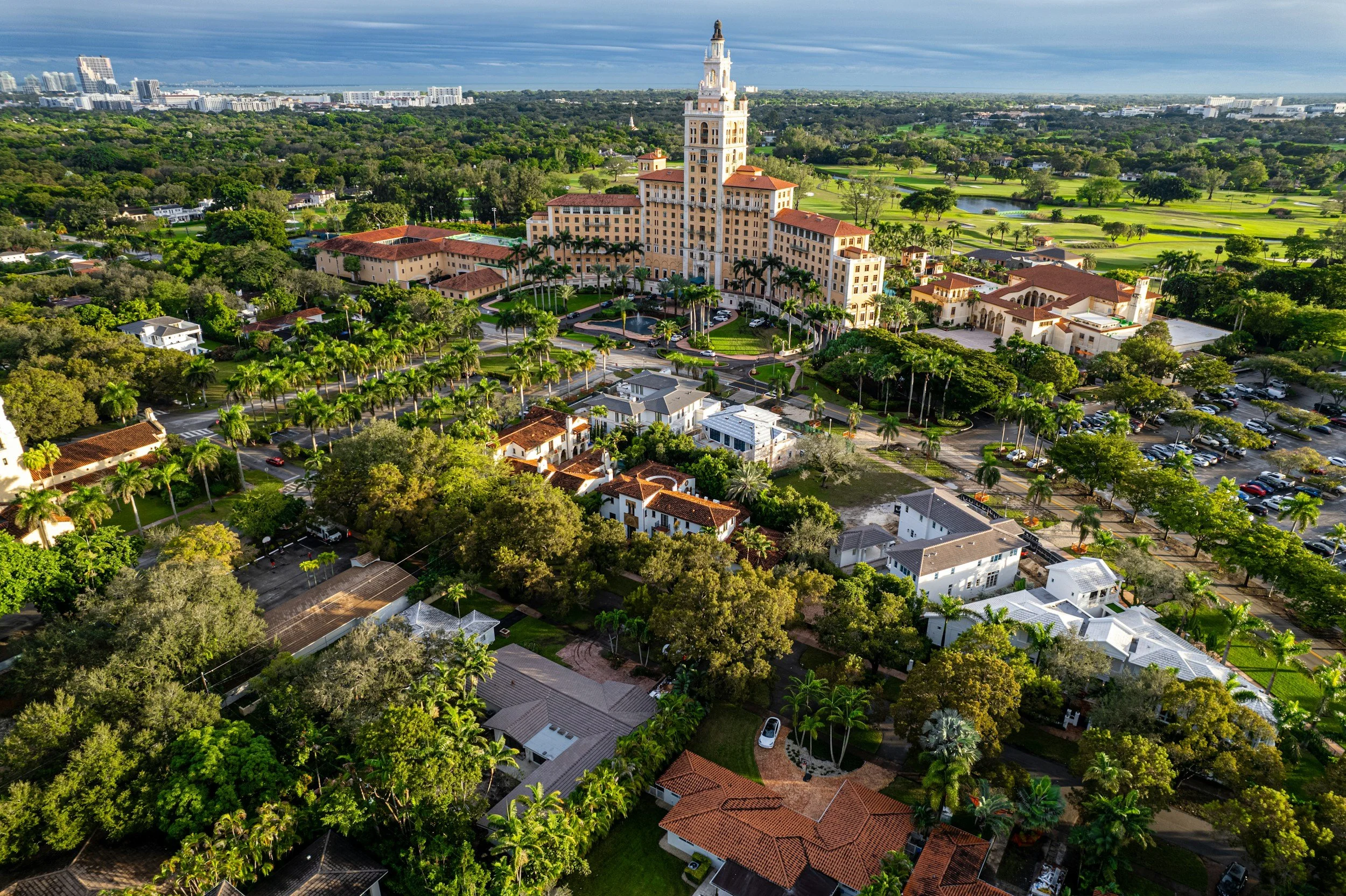 Aerial view of a large, historic, Mediterranean-style building with a tall central tower, surrounded by lush greenery, smaller residential houses, and parking lots, with a cityscape of high-rise buildings in the distance under a partly cloudy sky.